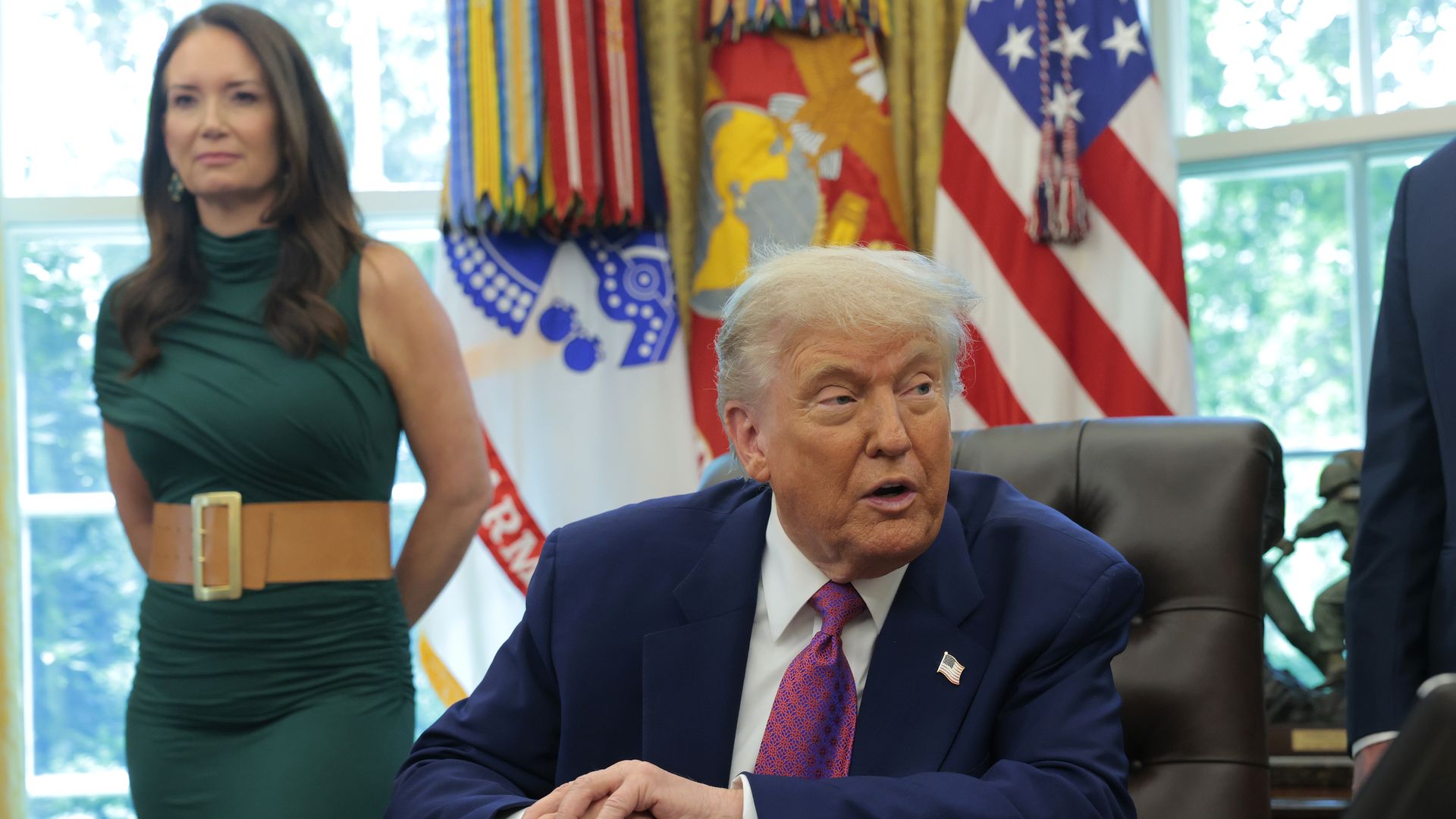 President Trump sits at a desk wearing a blue suit jacket, white shirt and red tie, while a woman stands behind him to his right wearing a sleeveless green dress.