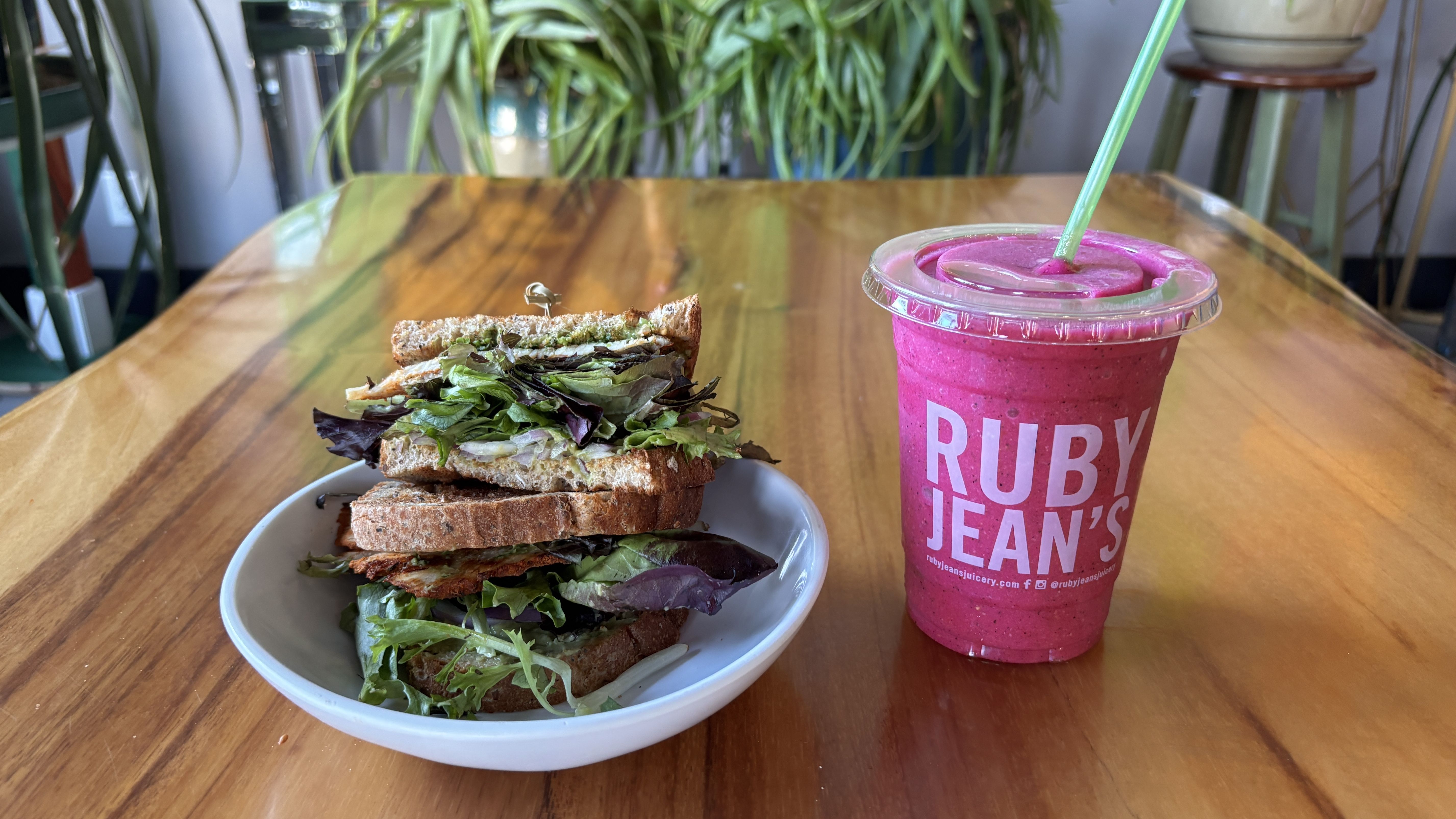 Sandwich with leafy greens on multigrain bread on a white plate next to a bright pink smoothie in a clear cup with green straw and "RUBY JEAN'S" logo on a wooden table.