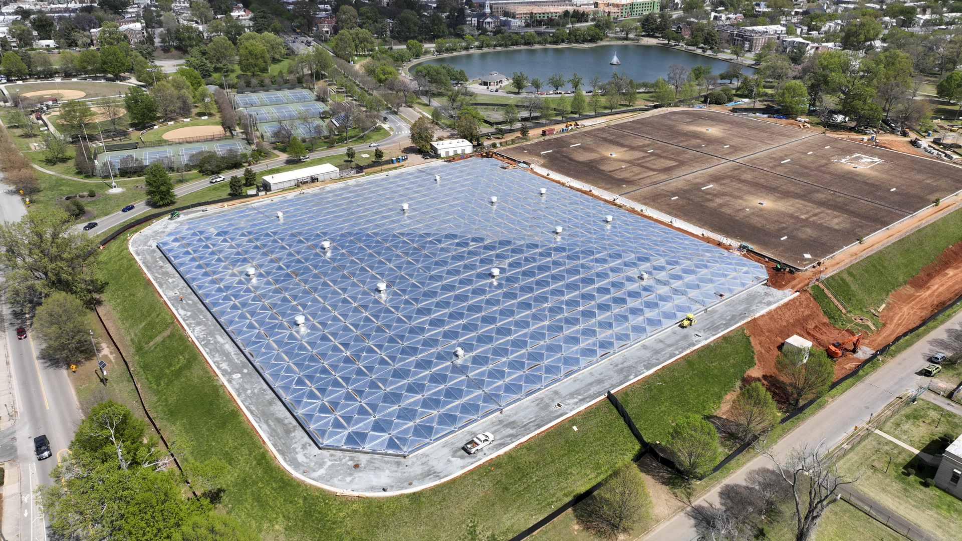 Aerial view of a construction site with a large glass-geodesic roof of triangular panels next to a dirt-covered field; nearby park, tennis courts, a lake, and urban area in distance.