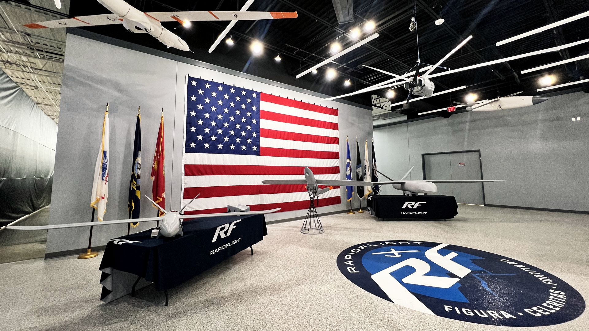 A well-lit showroom in a factory, displaying different size drones. A large American flag is seen against a back wall.