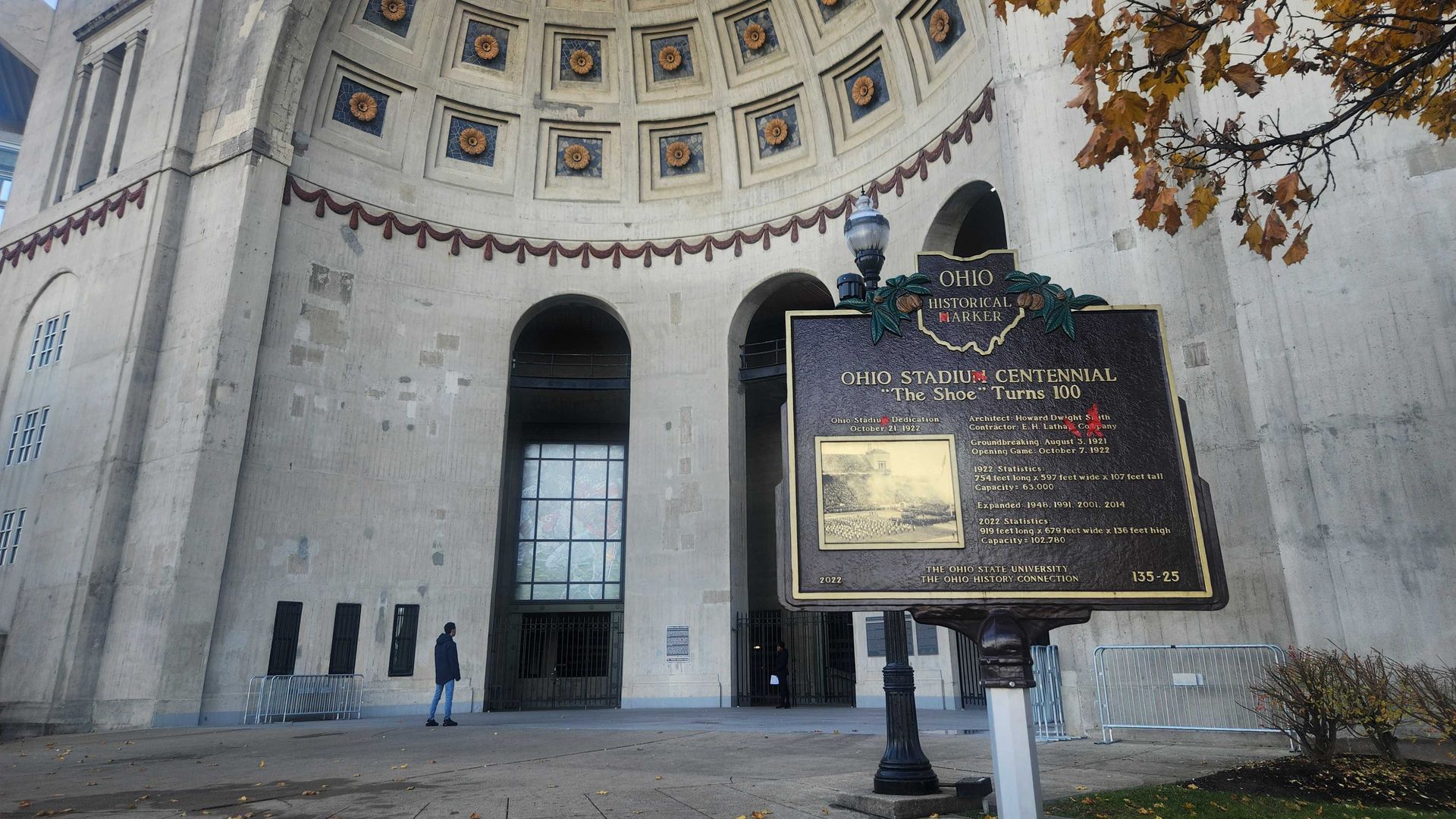 Exterior of Ohio Stadium with a large historical marker in front detailing the stadium's centennial, gray structure with decorative dome ceiling, and autumn trees with orange leaves nearby.