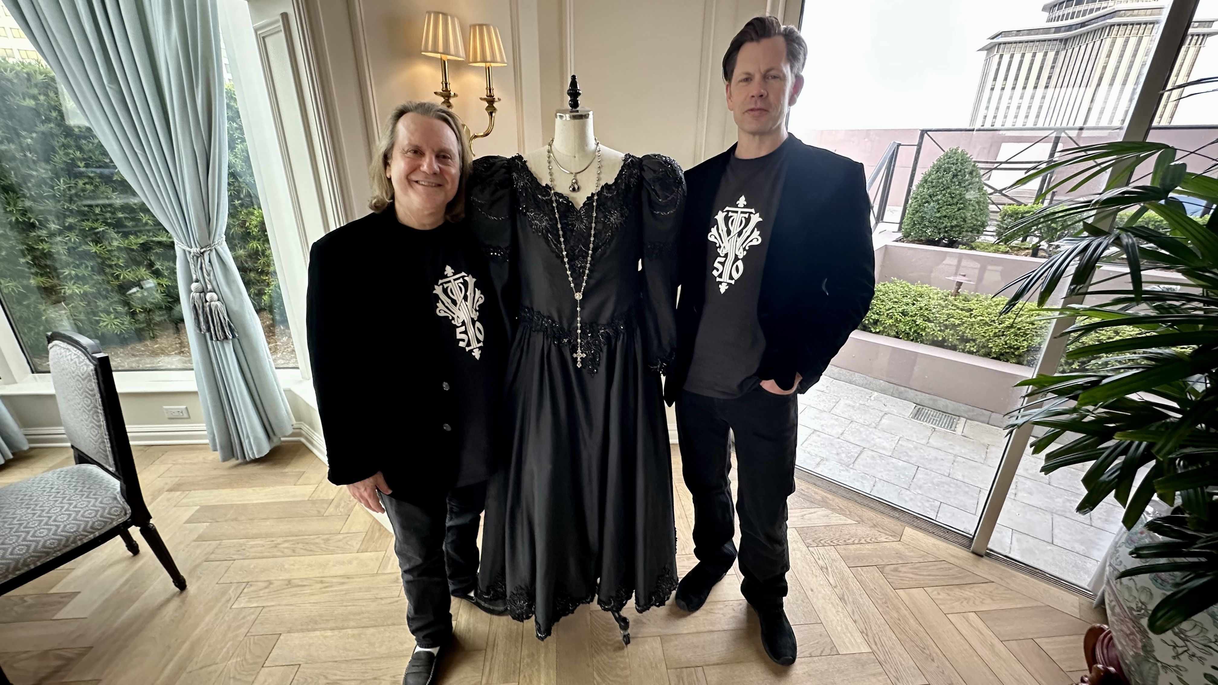 Three people pose indoors with a black lace gown on a dress form; two men in black outfits with a white crest logo flank the mannequin amid bright windows, blue curtains, and greenery.