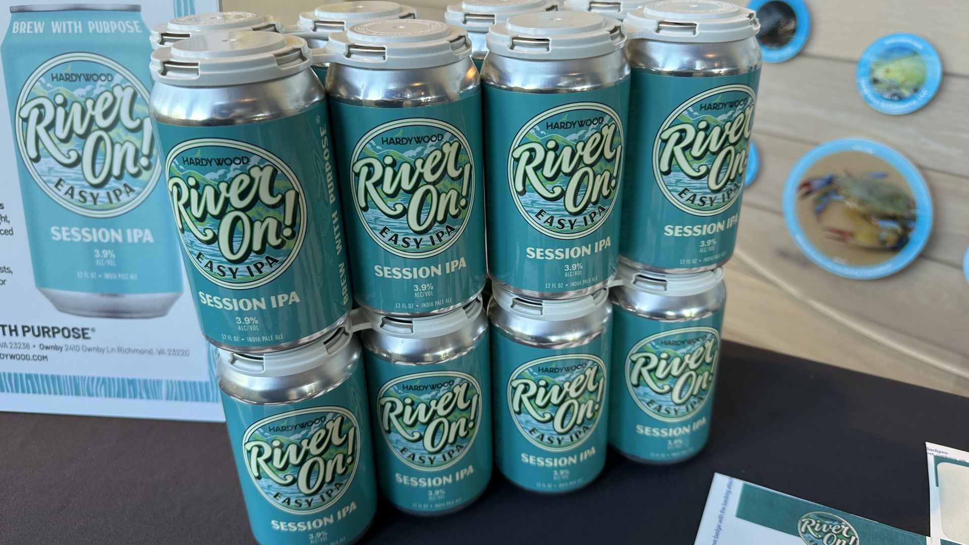 Teal River On! beer cans (Easy IPA and Session IPA) by Hardywood, stacked in front of a display box reading "Brew with Purpose"; blue circular plaques decorate a light wood wall in the background.