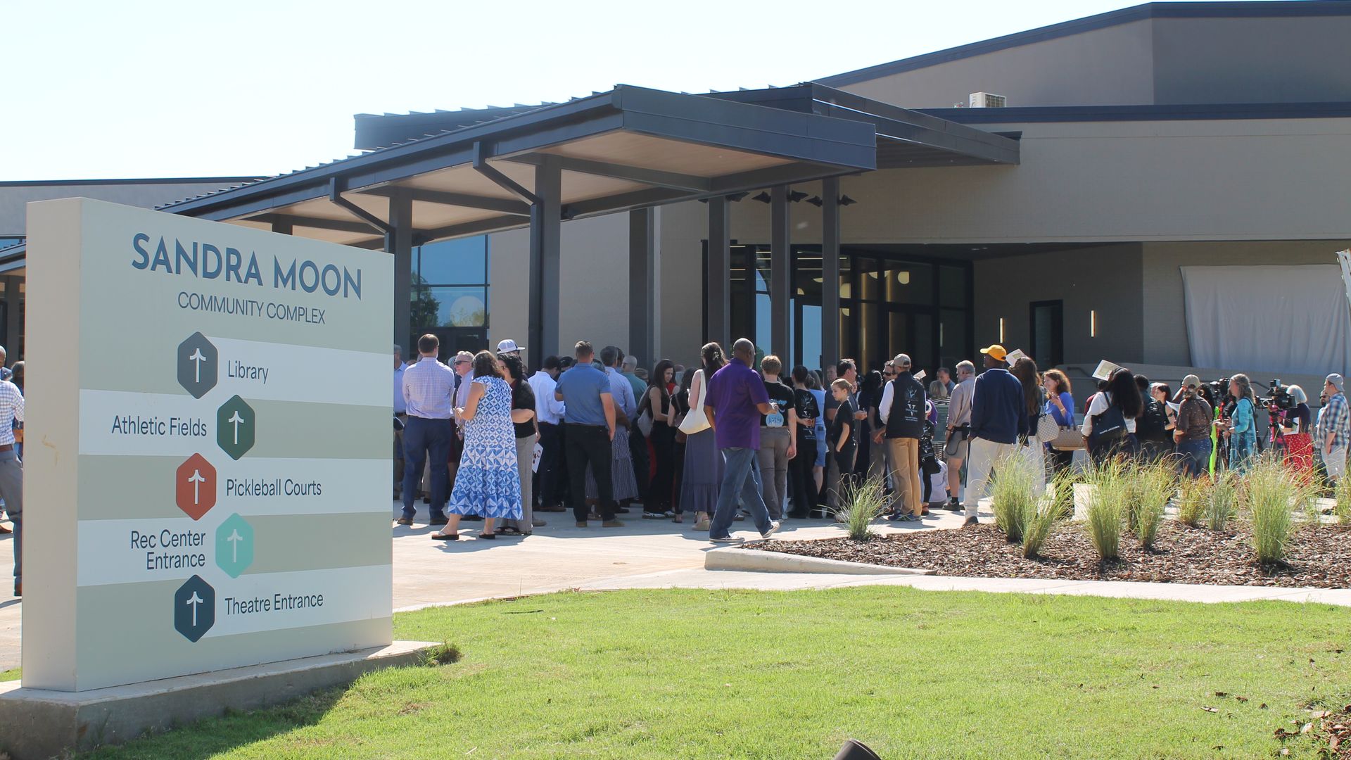 Crowd outdoors at the Sandra Moon Community Complex under a dark metal canopy. A sign reads the complex name and directs to Library, Athletic Fields, Pickleball Courts, Rec Center, and Theatre Entrance.