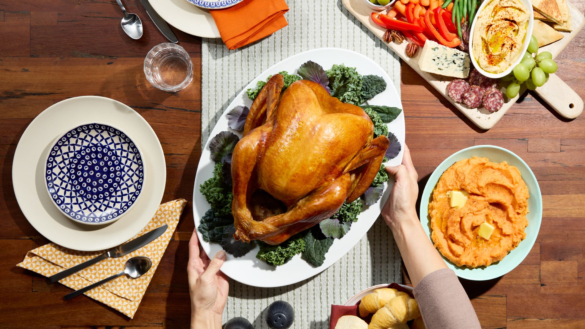 A photo showing a table set with a golden brown turkey on a platter garnished with greens. Sides of mashed potatoes and a charcuterie board are also visible. 