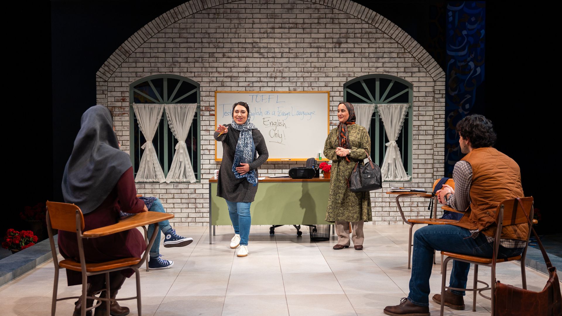 Classroom scene with two women standing near a desk; one speaks pointing forward. Three students sit at desks facing them. A whiteboard behind reads "TOEFL English Only."