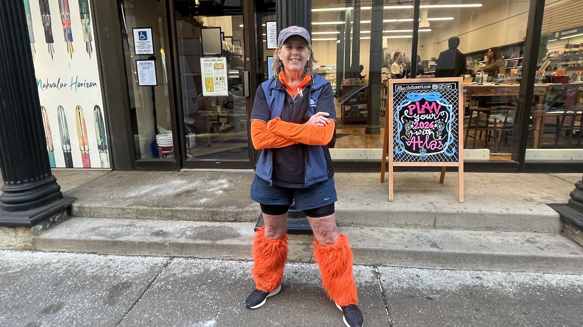 Smiling person wearing a navy cap, blue vest over black shirt and shorts, bright orange furry leg warmers, standing with arms crossed outside a store with a chalkboard sign saying "Plan your 2026 with Atlas".