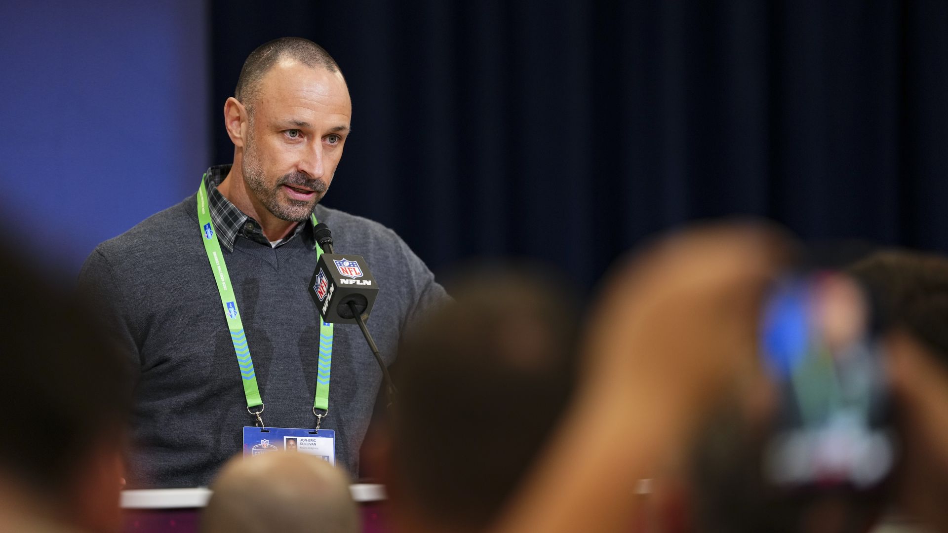 A man wearing a gray sweater over a plaid collared shirt stands at a podium speaking into a microphone while a crowd of reporters in the foreground watch, some recording with their phones. 