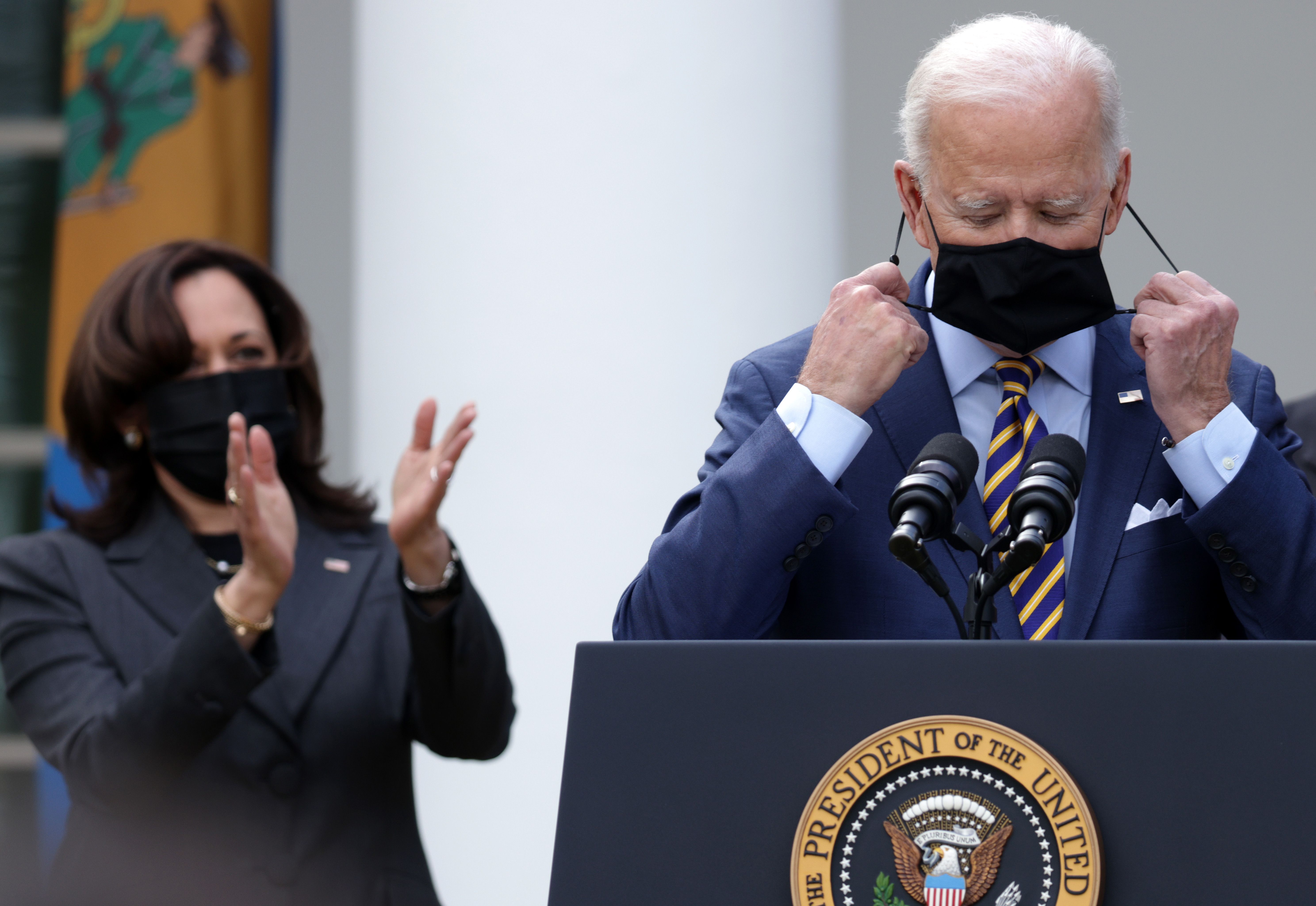 Biden takes off his mask at a podium, while Harris, in a mask, stands next to him and claps