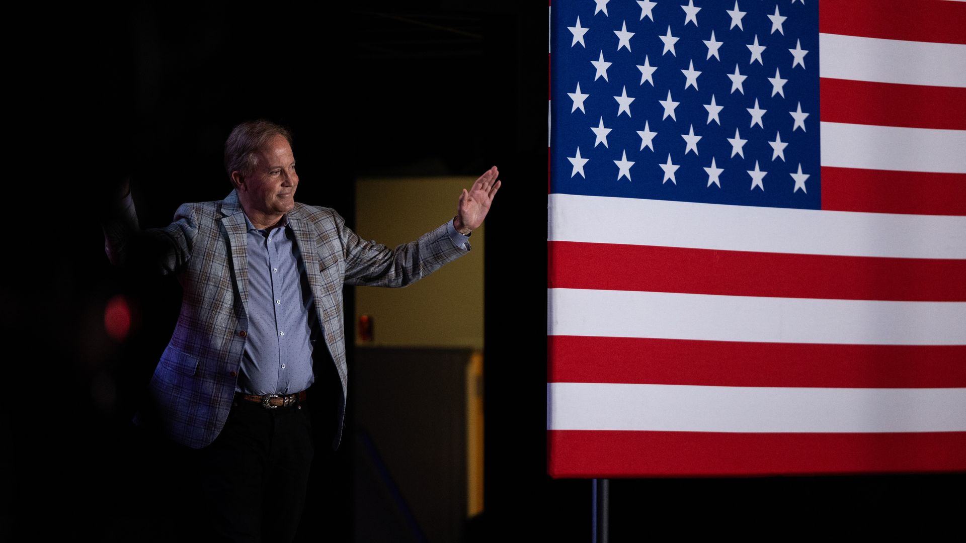 Ken Paxton waves at a crowd while standing behind a U.S> flag.