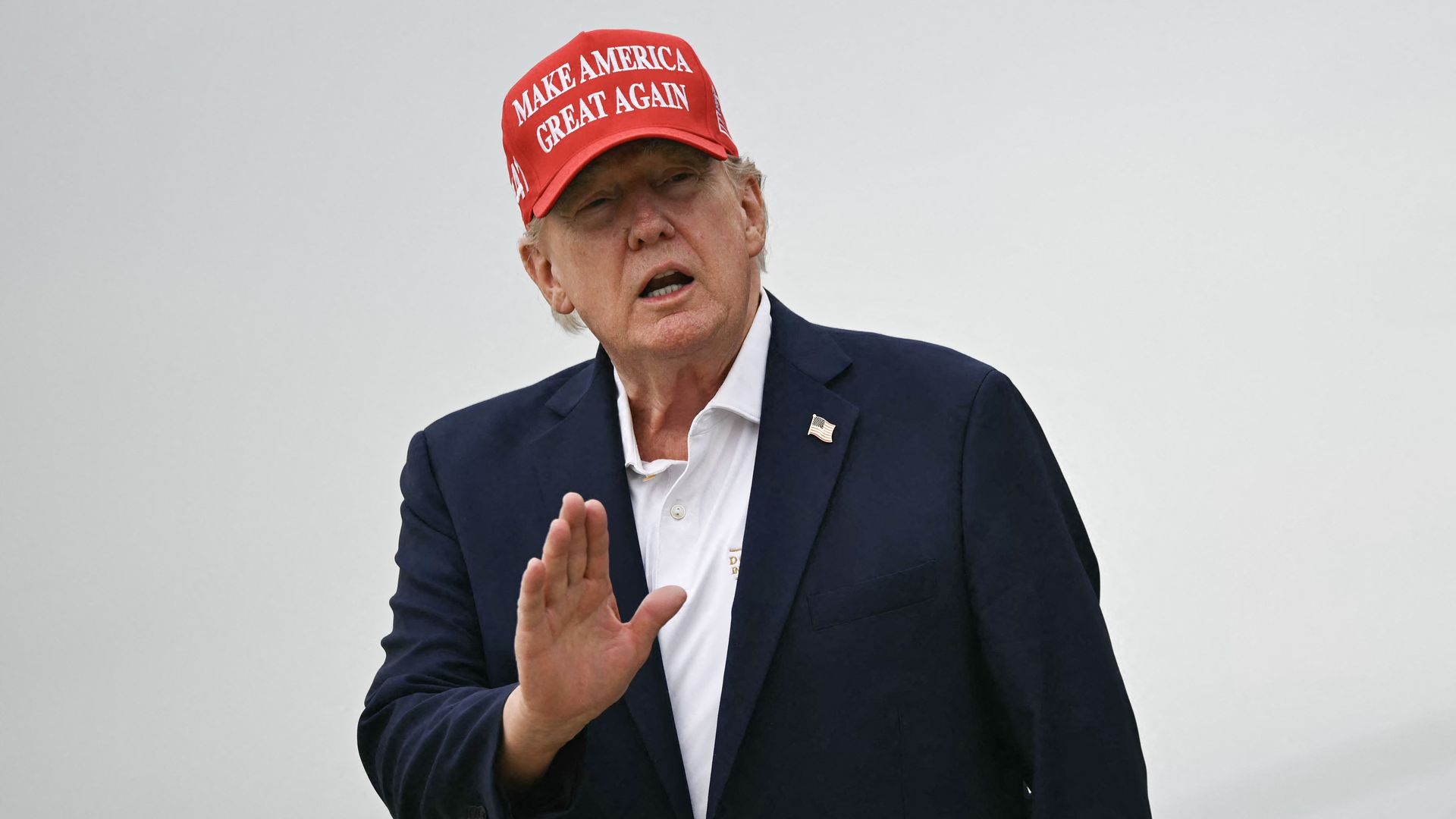 President Trump in a navy suit and white shirt wears a red cap reading MAKE AMERICA GREAT AGAIN and gestures with his hands against a plain gray background.