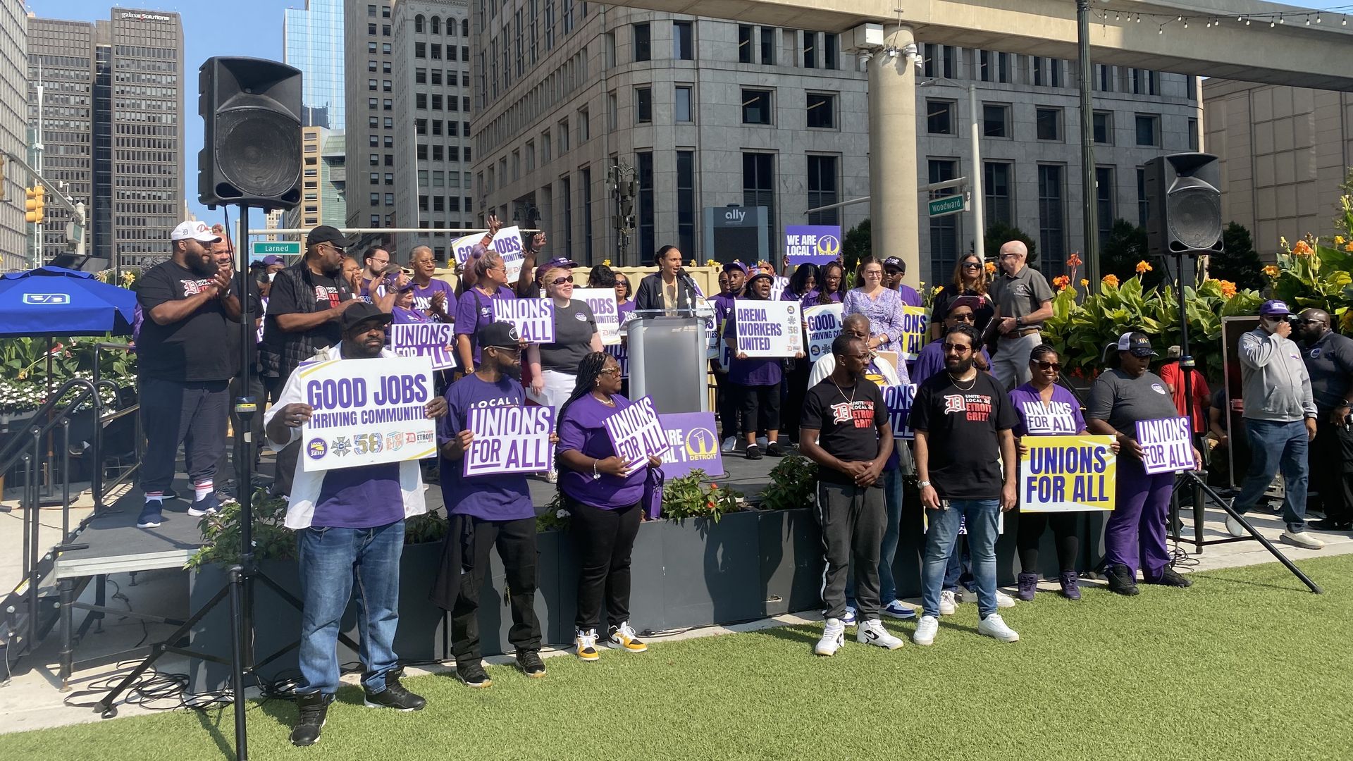 Mary Sheffield speaks in the center of a crowd of purple-clad people, in front of large downtown buildings.