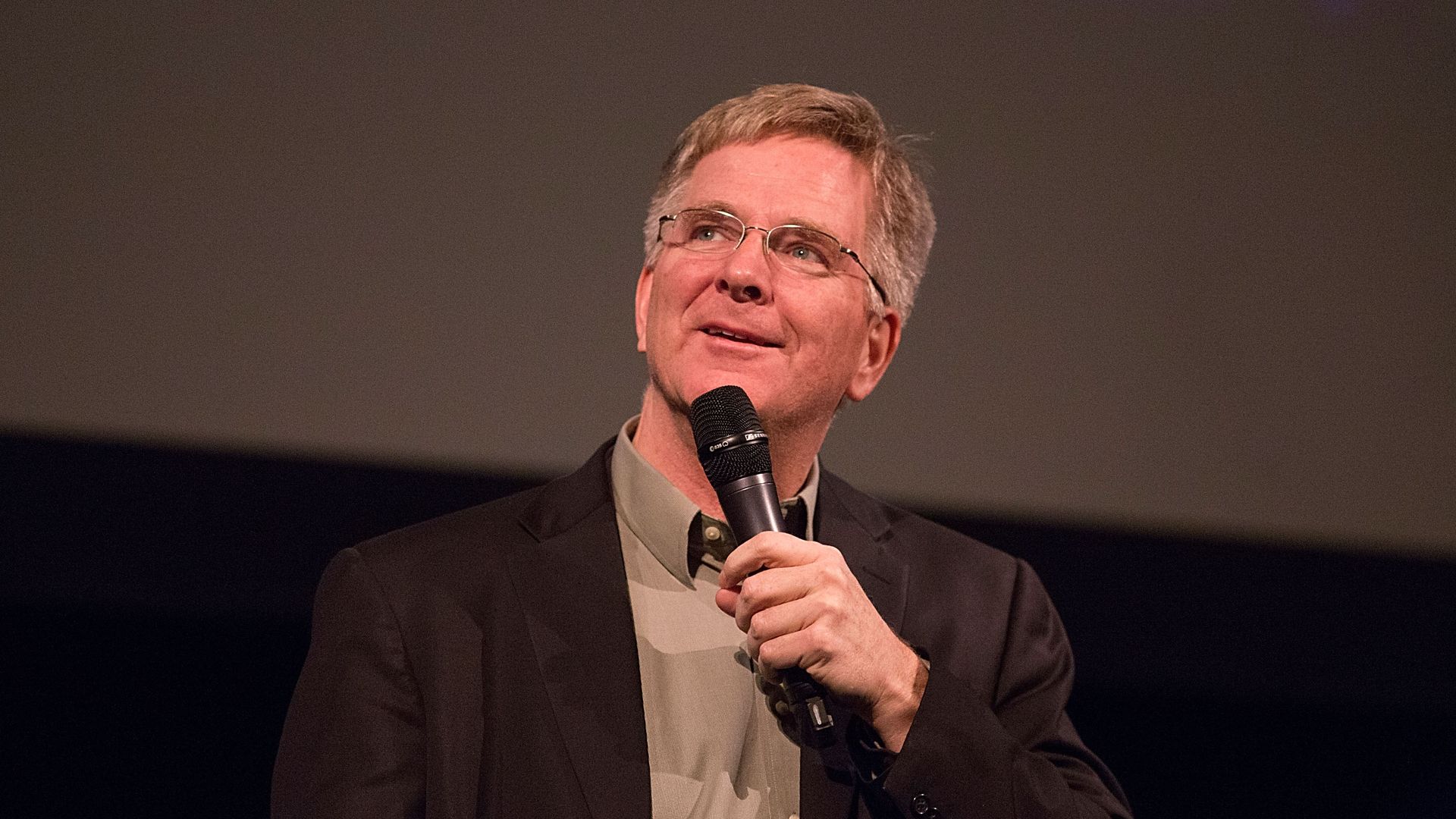 Middle-aged man with glasses, gray hair, and a beard, wearing a dark suit and light shirt, holding a microphone and looking upward while speaking on stage.