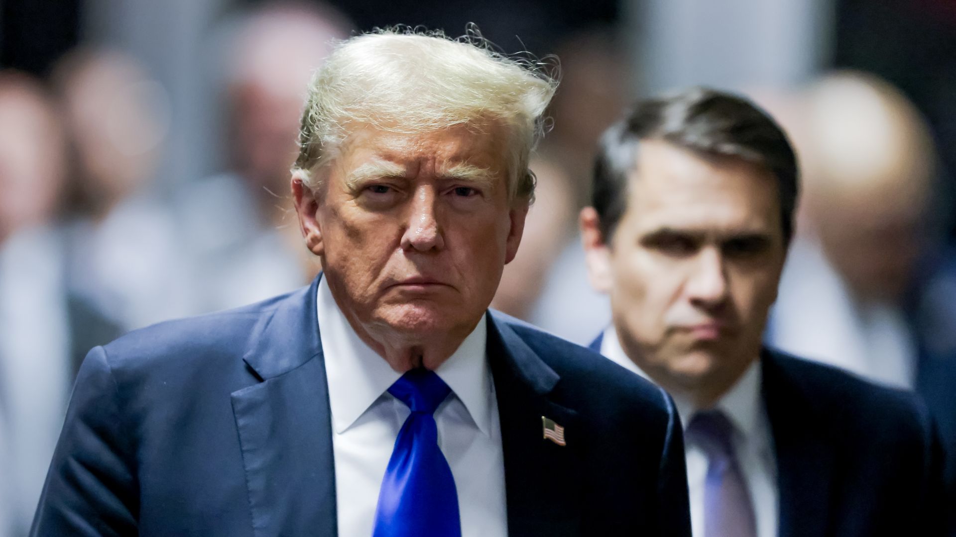 President Trump — wearing a dark suit, a white collared shirt, a blue tie and an American flag pin — walks with Todd Blanche — wearing a dark suit, white collared shirt and tie — in the background. 