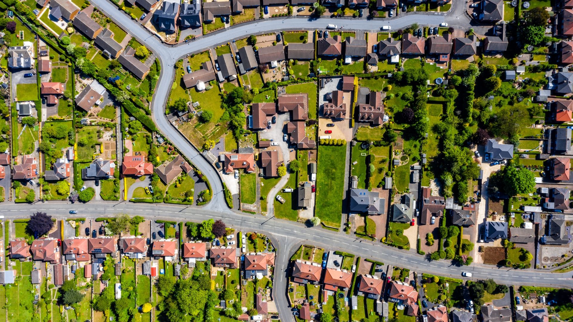 Aerial view of a suburban neighborhood with houses, green lawns, trees, and intersecting streets under clear sunlight.