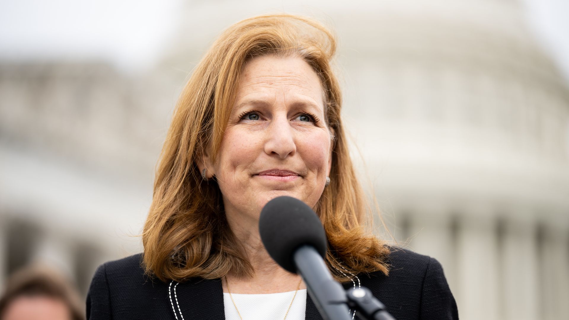 A woman stands outside at a microphone wearing dark suit jacket as wind blows her hair slightly.