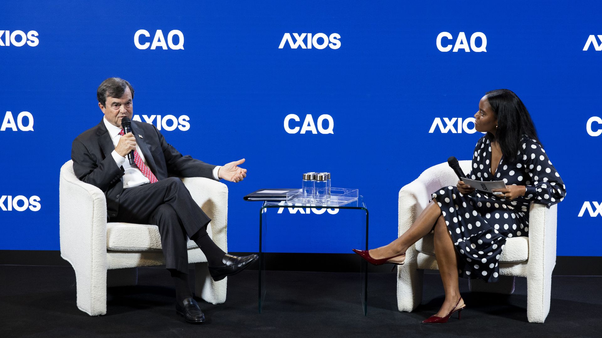 Two people sit in white armchairs on stage with backdrop reading "AXIOS" and "CAQ." A man speaks into a microphone while a woman in polka-dot dress listens; a glass table sits between them.