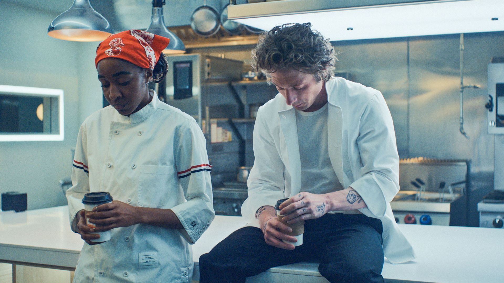 A photo showing two people sitting in a industrial-looking kitchen wearing white coats staring down at their coffees.