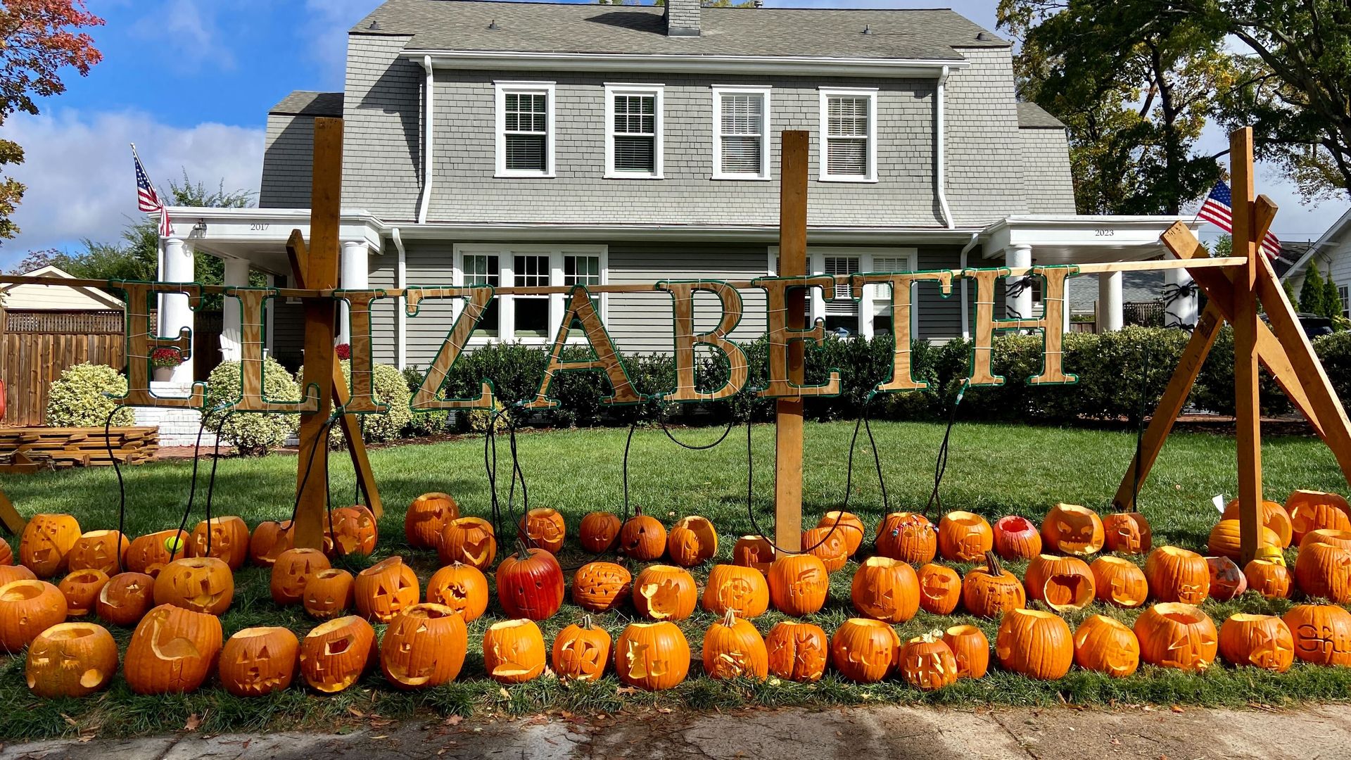 Large lawn display with the name ELIZABETH spelled out in large wooden letters above many carved pumpkins, in front of a gray two-story house with American flags.