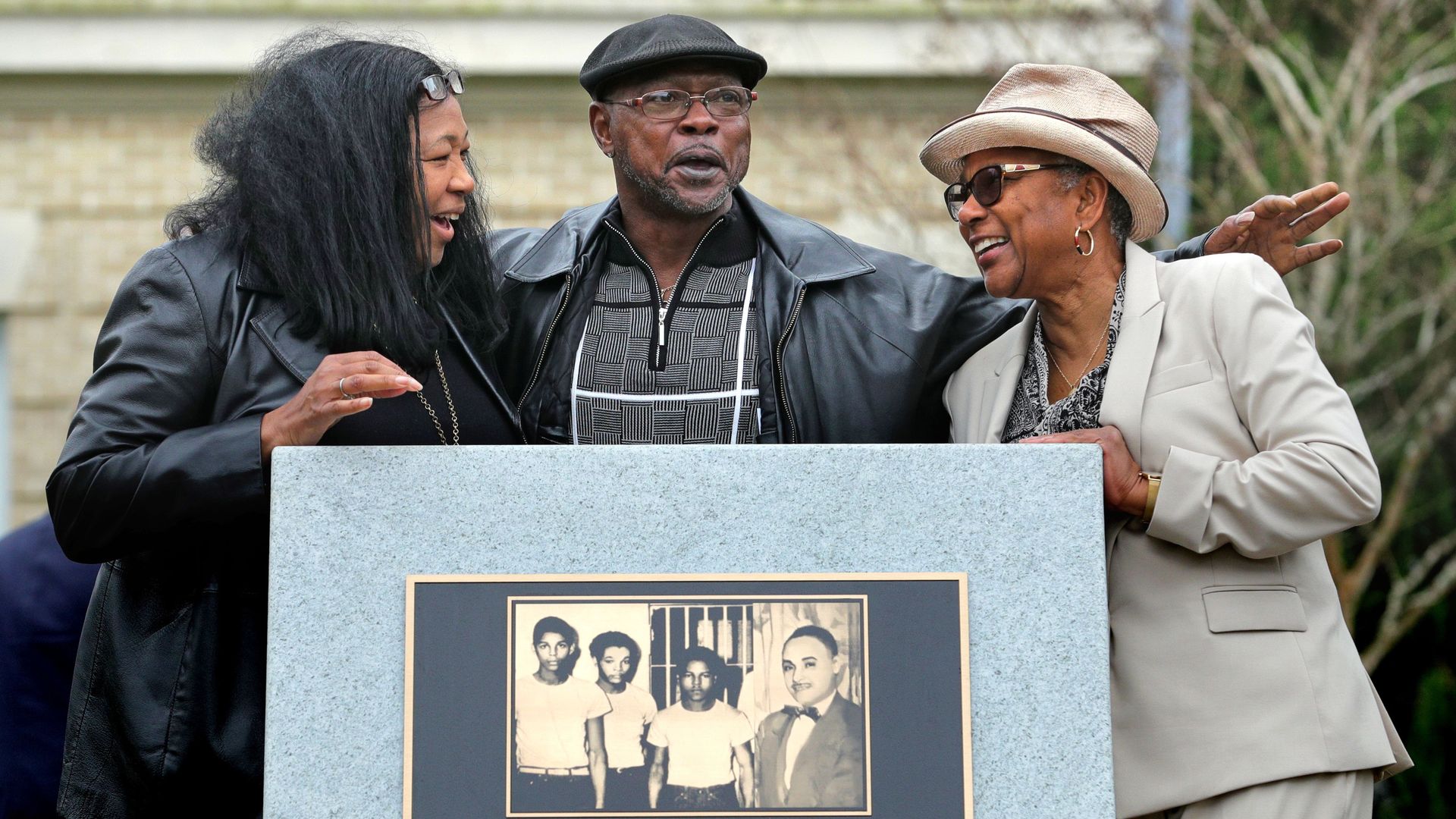 A man and two woman stand behind a granite monument with a plaque that reads "In Memory of the Groveland Four" under an image of four young men.