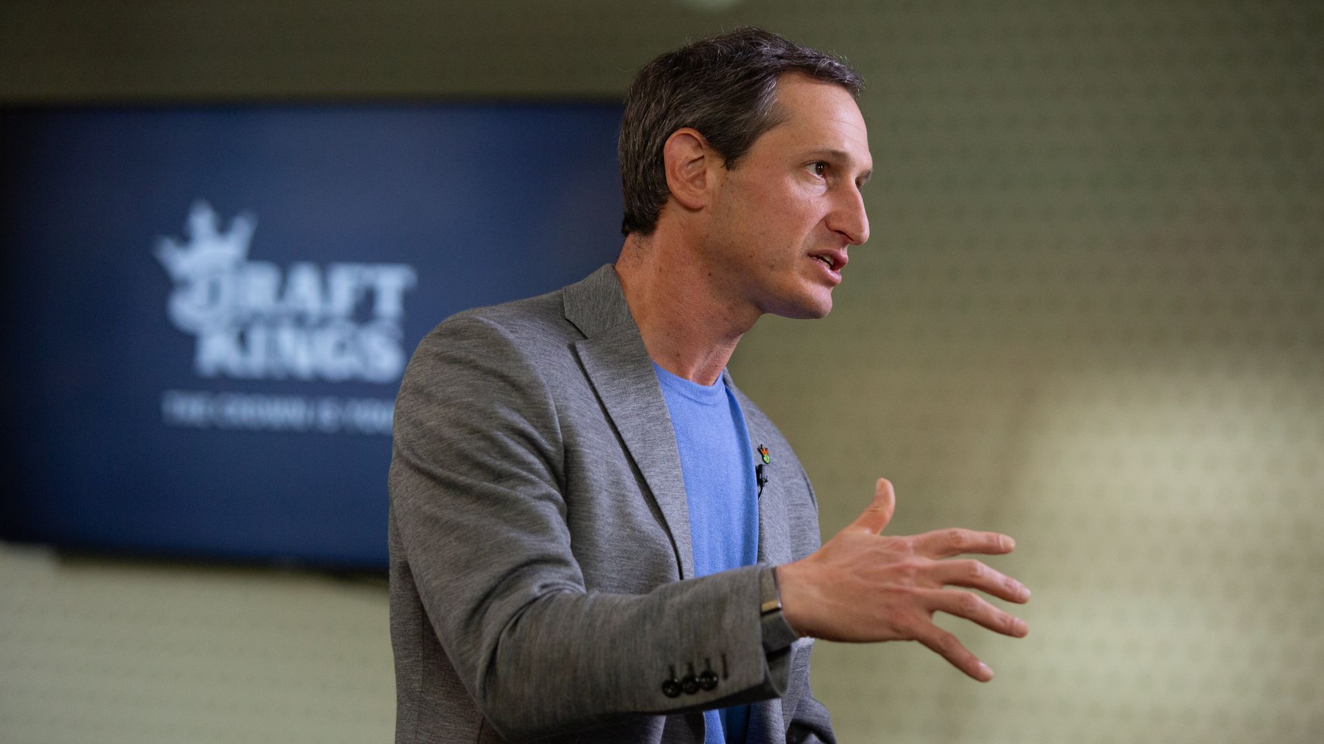 A person in a gray suit jacket and blue shirt speaks in front of a TV screen with the DraftKings logo