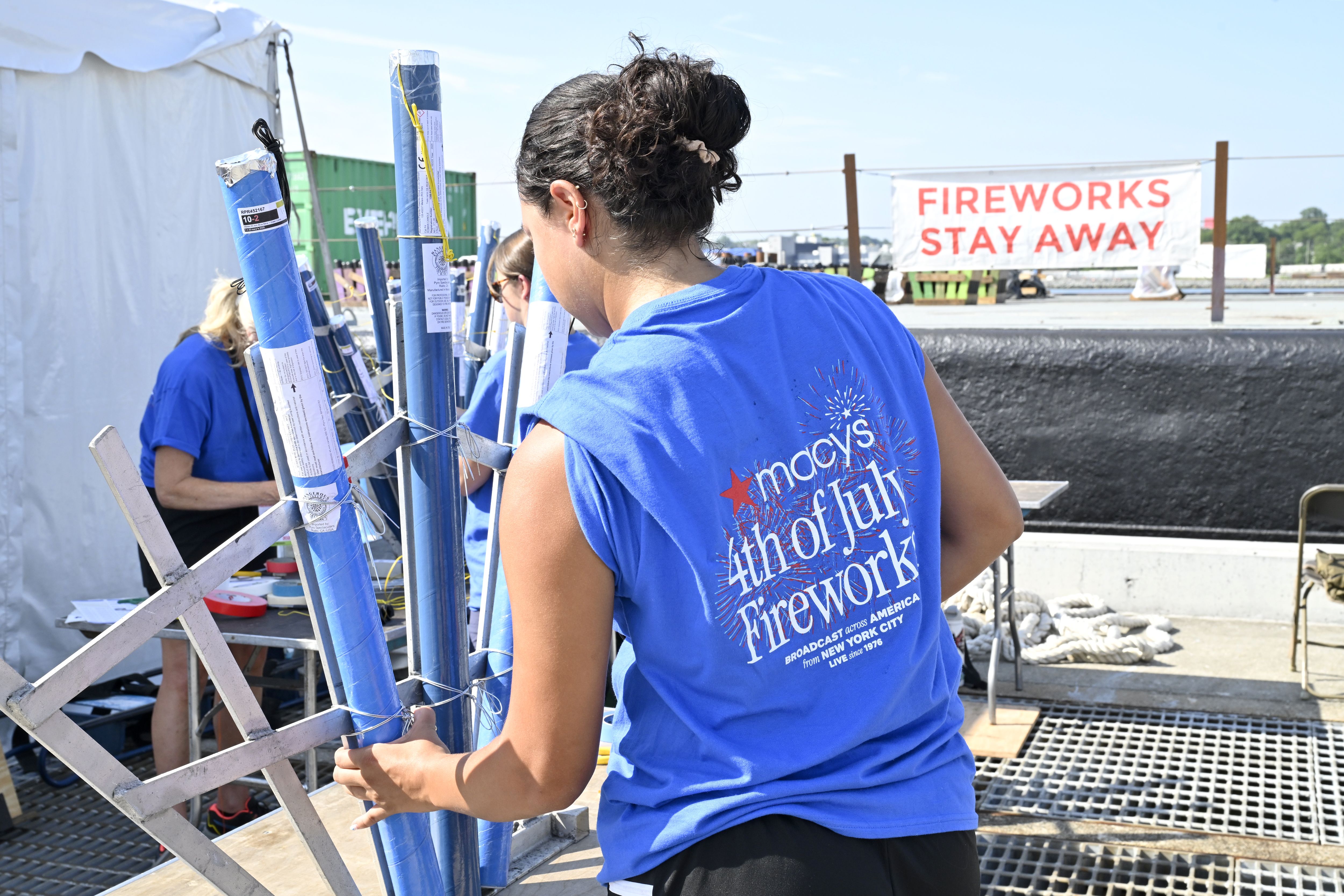 Person in a blue "Macy's 4th of July Fireworks" shirt handling large blue fireworks tubes, with a white sign reading "FIREWORKS STAY AWAY" in the background on a sunny day.