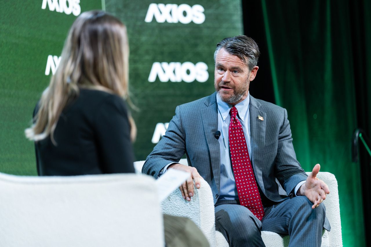 Man in gray suit and red tie speaking during an interview with a woman in a black jacket at an Axios event, with green backdrop and "AXIOS" logos visible.