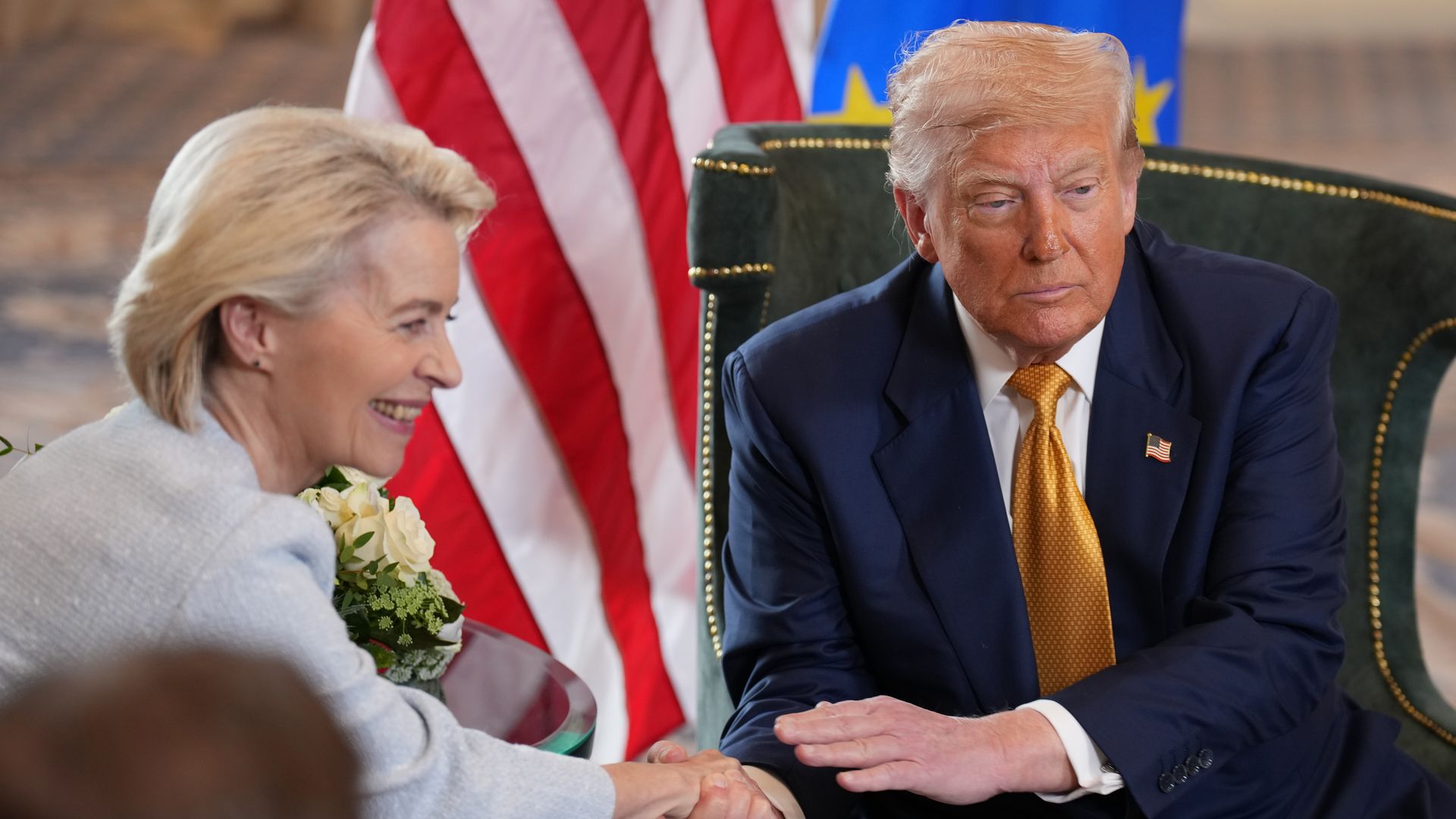 A woman in a light gray jacket smiles while shaking hands with a man in a navy suit and gold tie, sitting with American and European Union flags behind them.