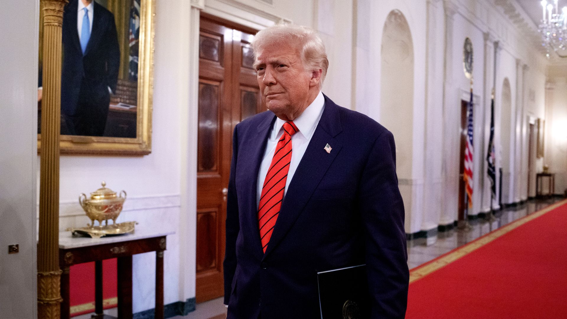 Trump walks toward the East Room of the White House with a folder under his arm.