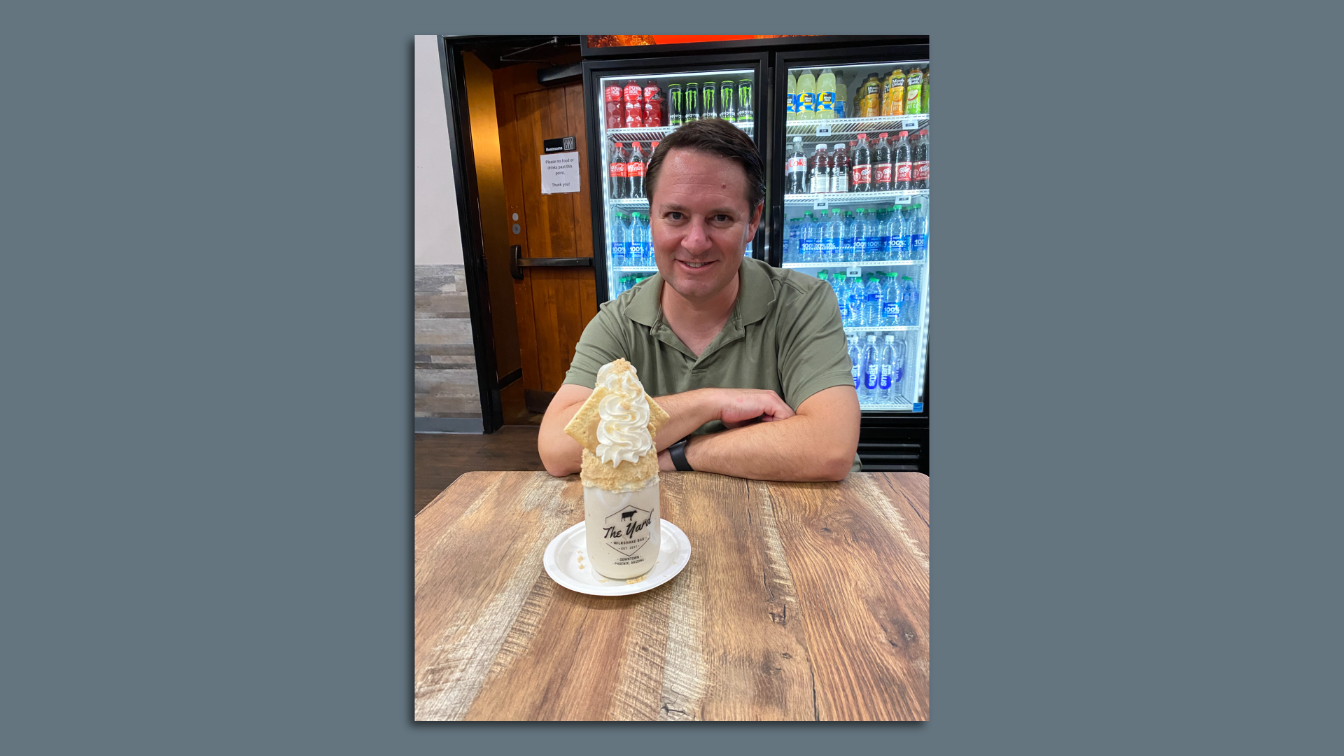 A man at a table with a white milkshake in a clear glass jar topped with whipped cream. 