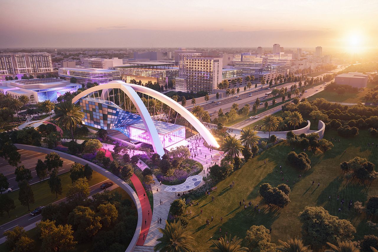 Aerial view of a modern cityscape at sunset with a unique building featuring two illuminated white arches and a colorful facade, surrounded by palm trees, green spaces, and roads.