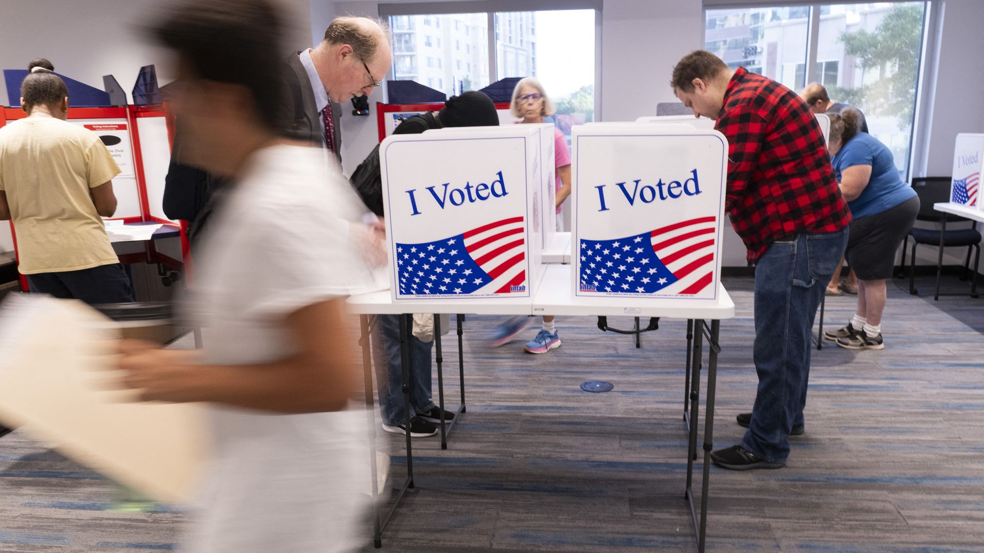 people voting in stations that say "I Voted" on the side. One person is blurred and holding a folder. 