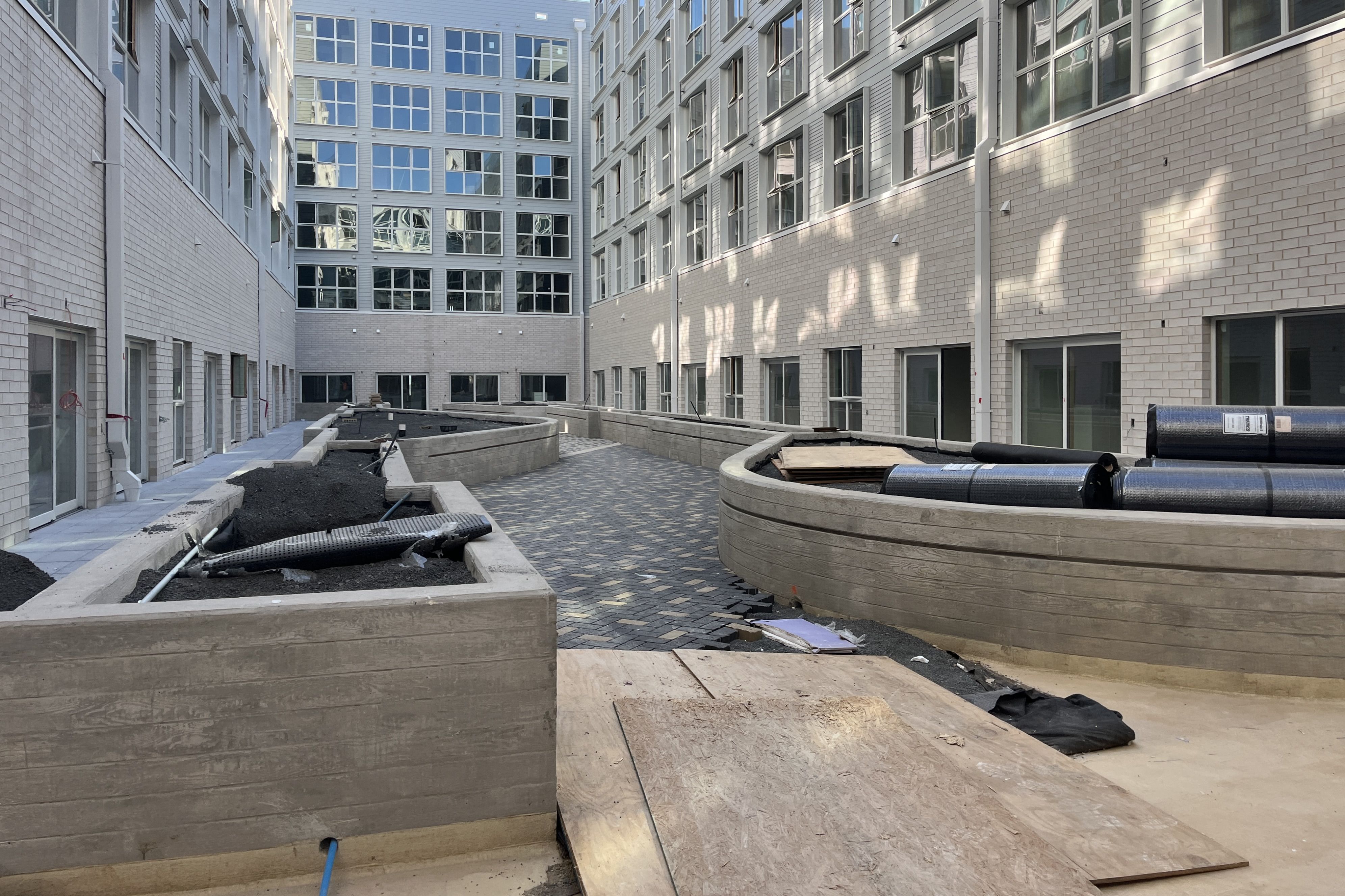 Construction site in a modern grey-brick apartment complex courtyard. Many windows, curved benches, and scattered pipes and debris on the paved ground.