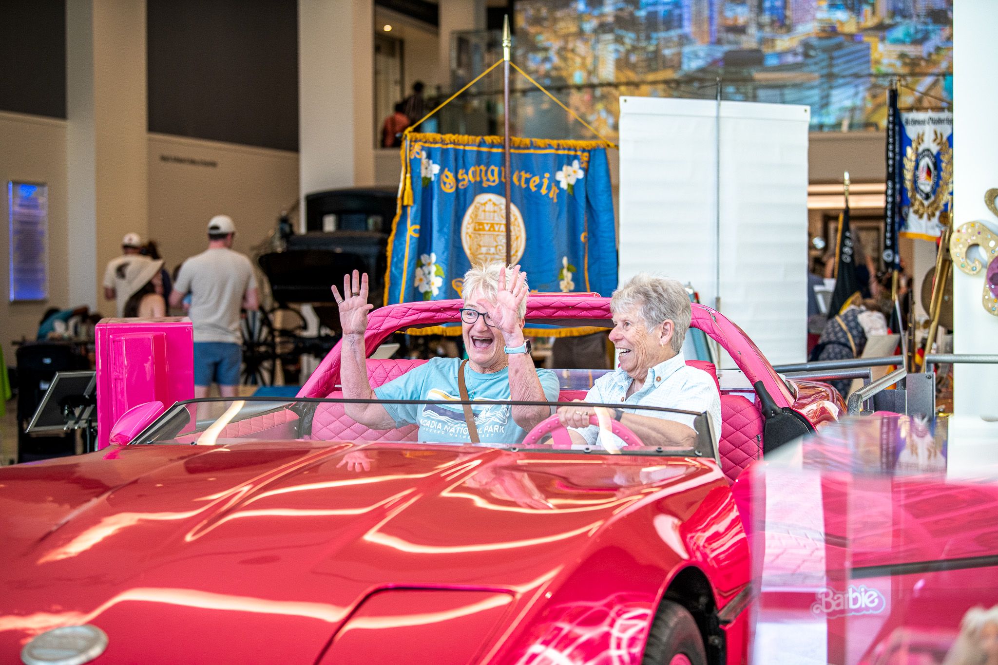 Two elderly women ride in a bright pink vintage convertible inside a busy exhibit hall, smiling and waving. A blue banner with floral designs and a cityscape mural looms behind them.