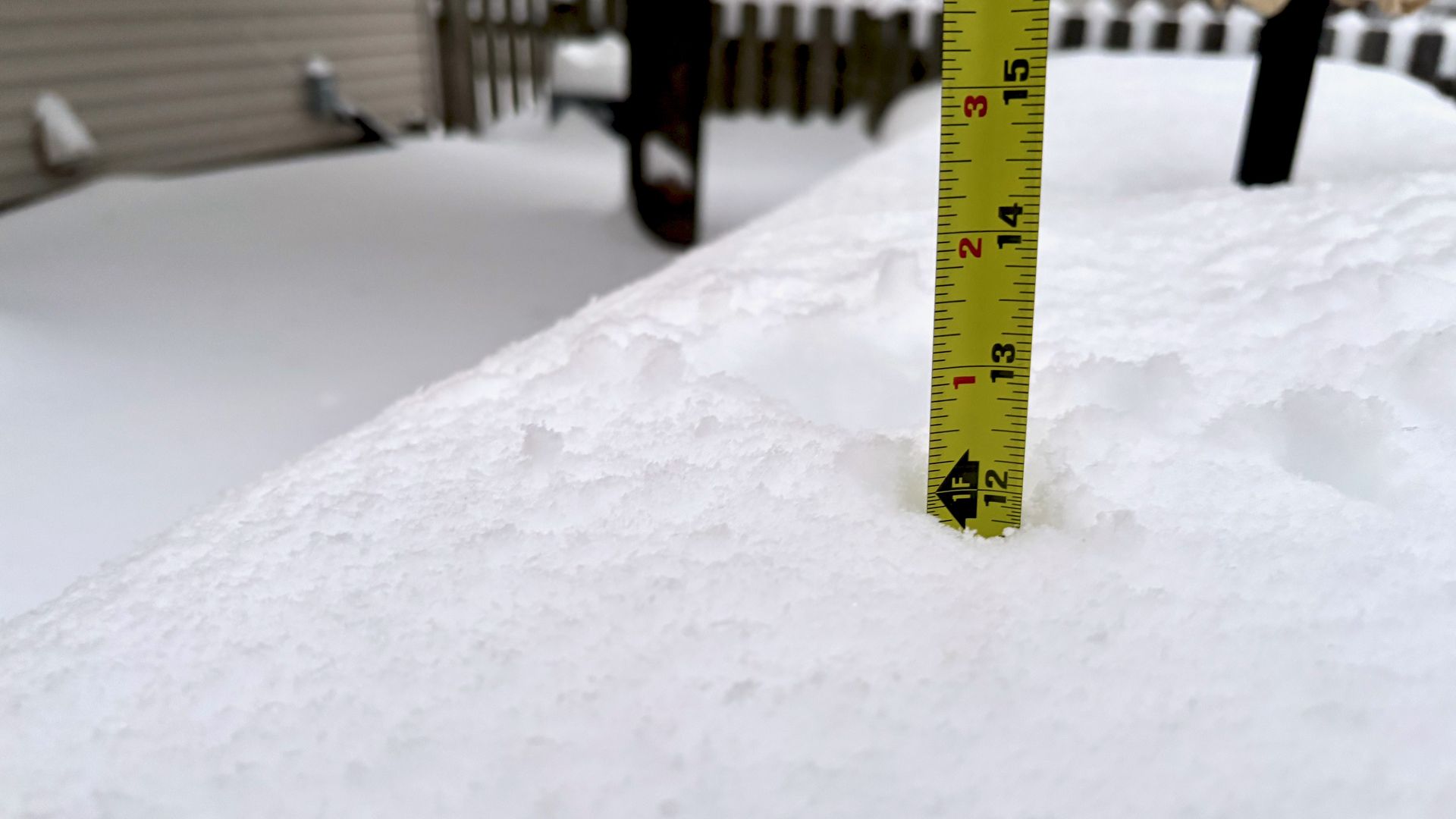 Yellow measuring tape inserted vertically into a snow-covered surface, showing snow depth of about 12 inches, with a blurred house and fence in the background.