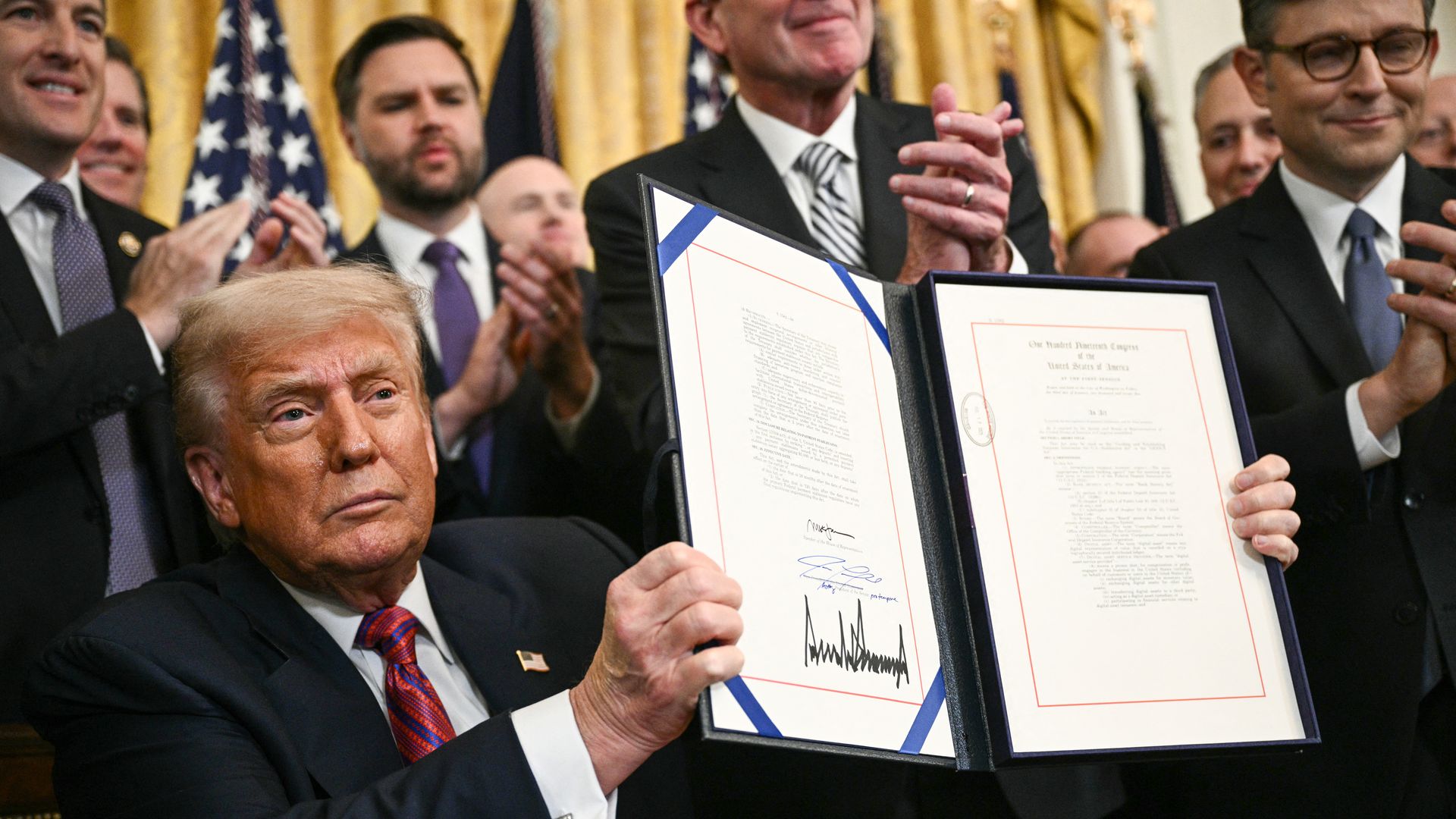 Former President Donald Trump in a dark suit and red tie holds up a signed official document while people in suits clap behind him, with American flags in the background.