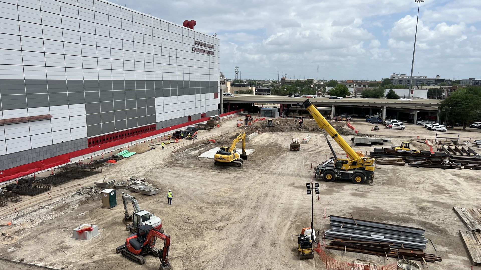 Construction site beside a white-gray building; yellow crane, excavator, stacked steel beams, and workers in safety vests operate on dirt near an overpass under a cloudy sky.