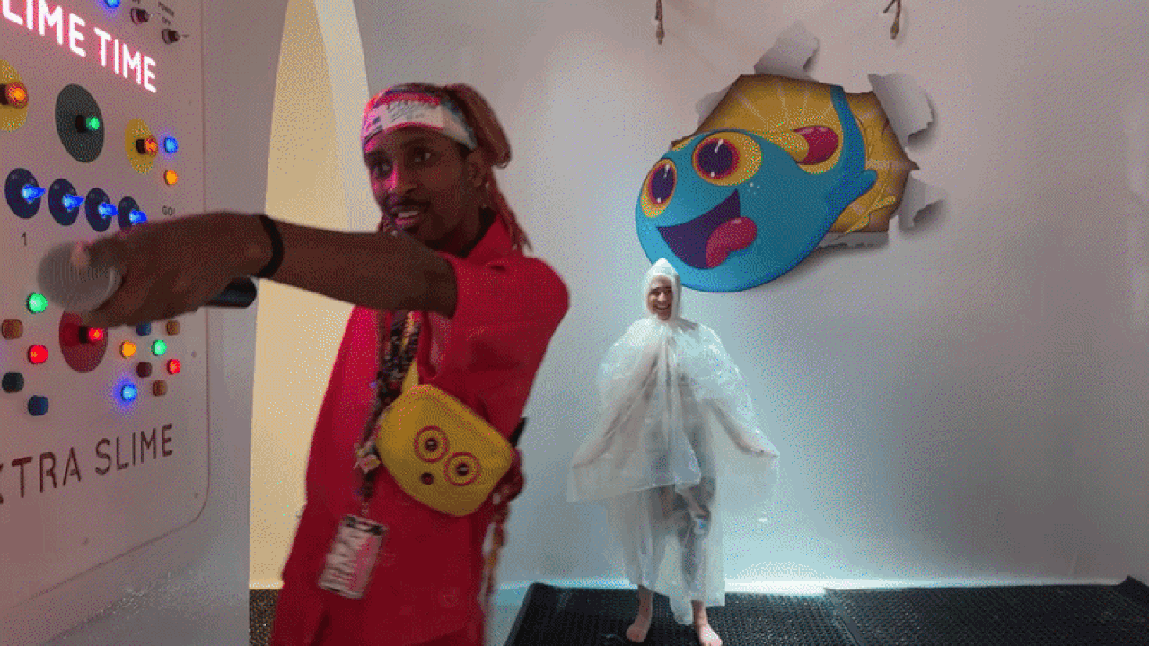 Man in red with a yellow emoji bag points at a wall panel with multicolored lights; a person in a translucent poncho stands near a blue cartoon slime mural on the wall.