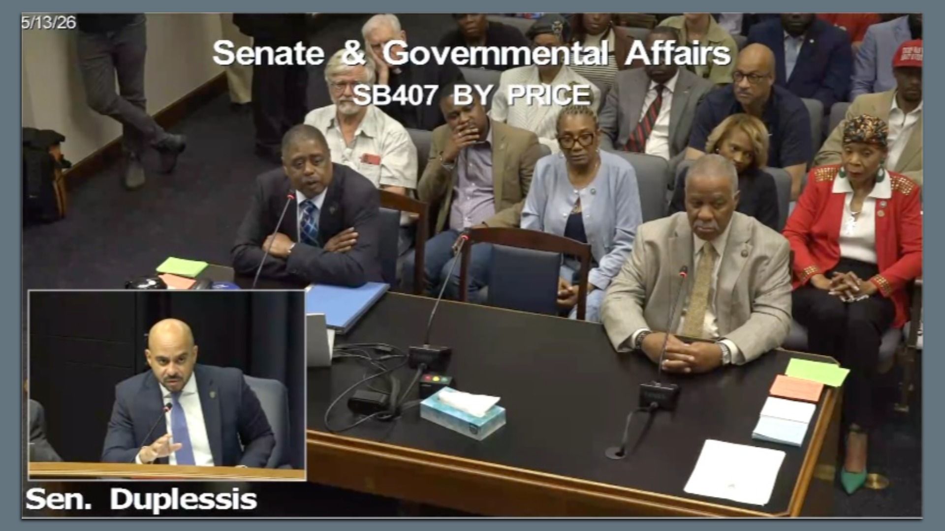 Hearing room with officials in suits seated; a man in a dark suit speaks into a microphone at a large desk, while an inset shows another man. Overlay reads \"Senate & Governmental Affairs\".