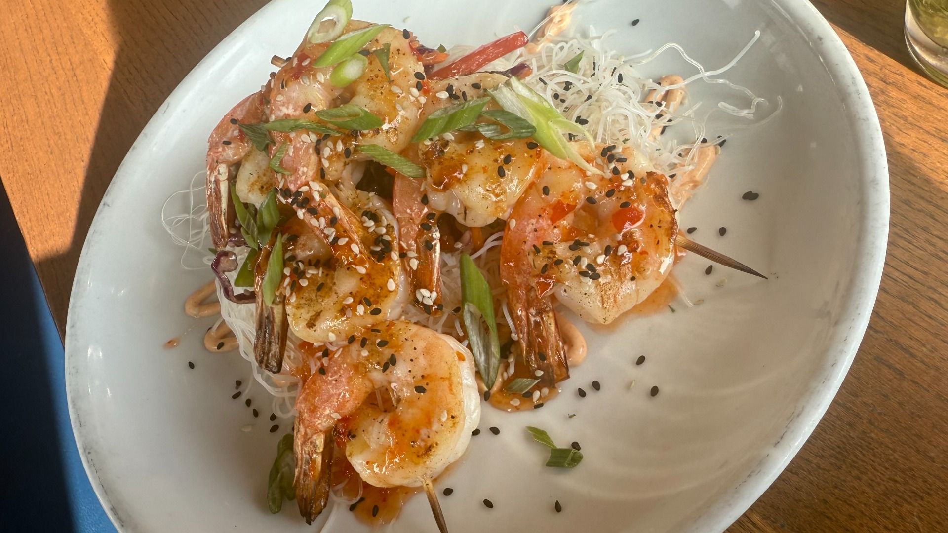 Grilled shrimp skewers glazed with orange sauce, garnished with black and white sesame seeds and green onions, served on a bed of white rice noodles on a white plate.