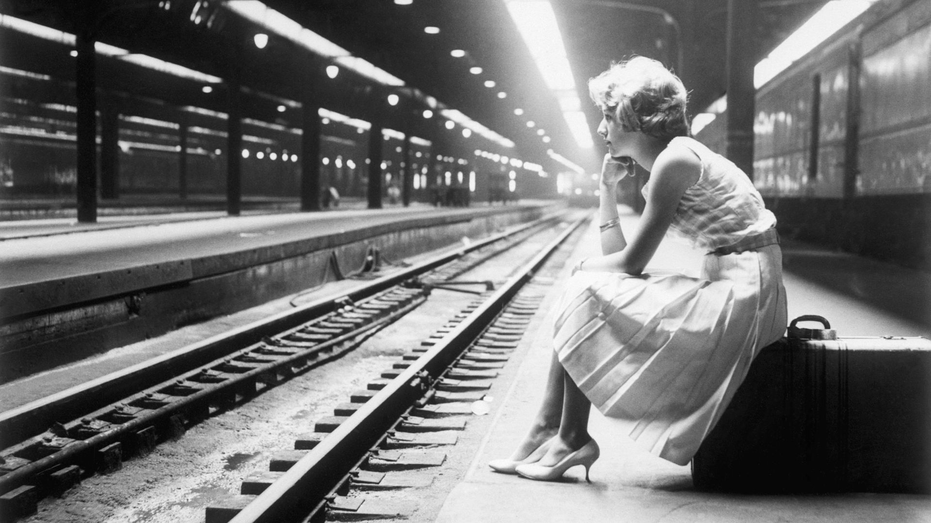 Black and white photo of a woman sitting on a suitcase on a train platform
