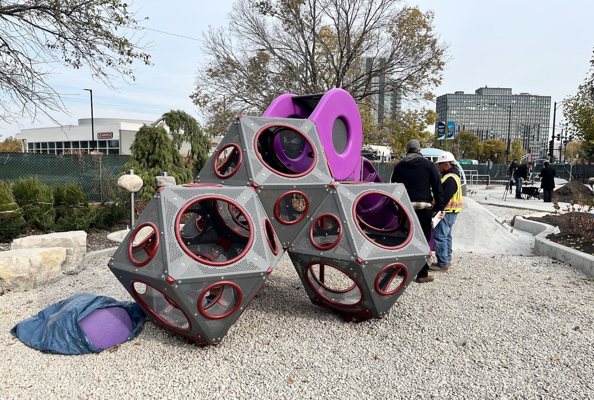 Geometric sculpture made of gray perforated metal cubes with purple rings sits on a gravel site; workers in hard hats stand nearby as trees and city buildings loom in the background.