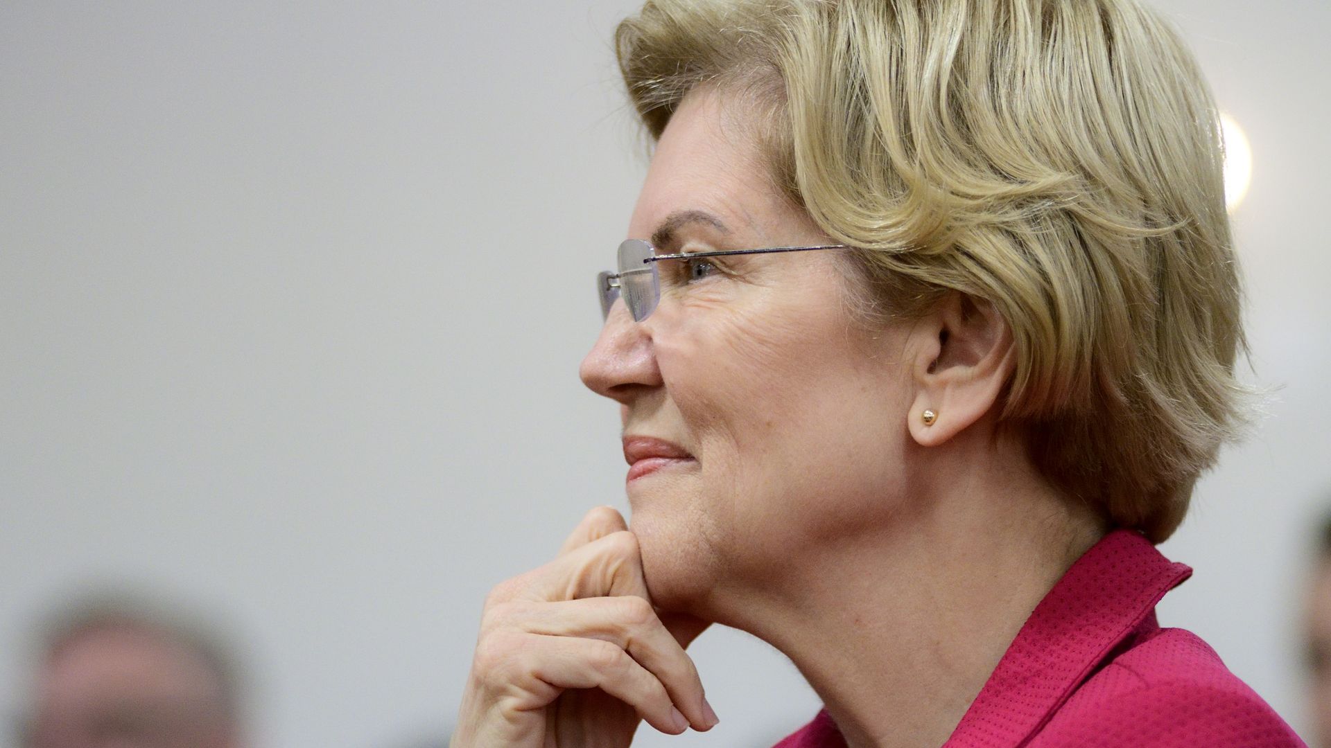 Warren smiling while touching her hand to her face. She is wearing a pink blazer-looking jacket and glasses. 