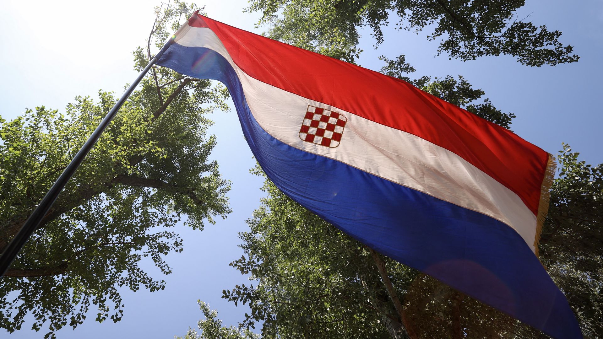 Croatian flag waving on a flagpole against a clear blue sky, with green trees around; red, white, blue horizontal stripes and the checkered crest in the center.
