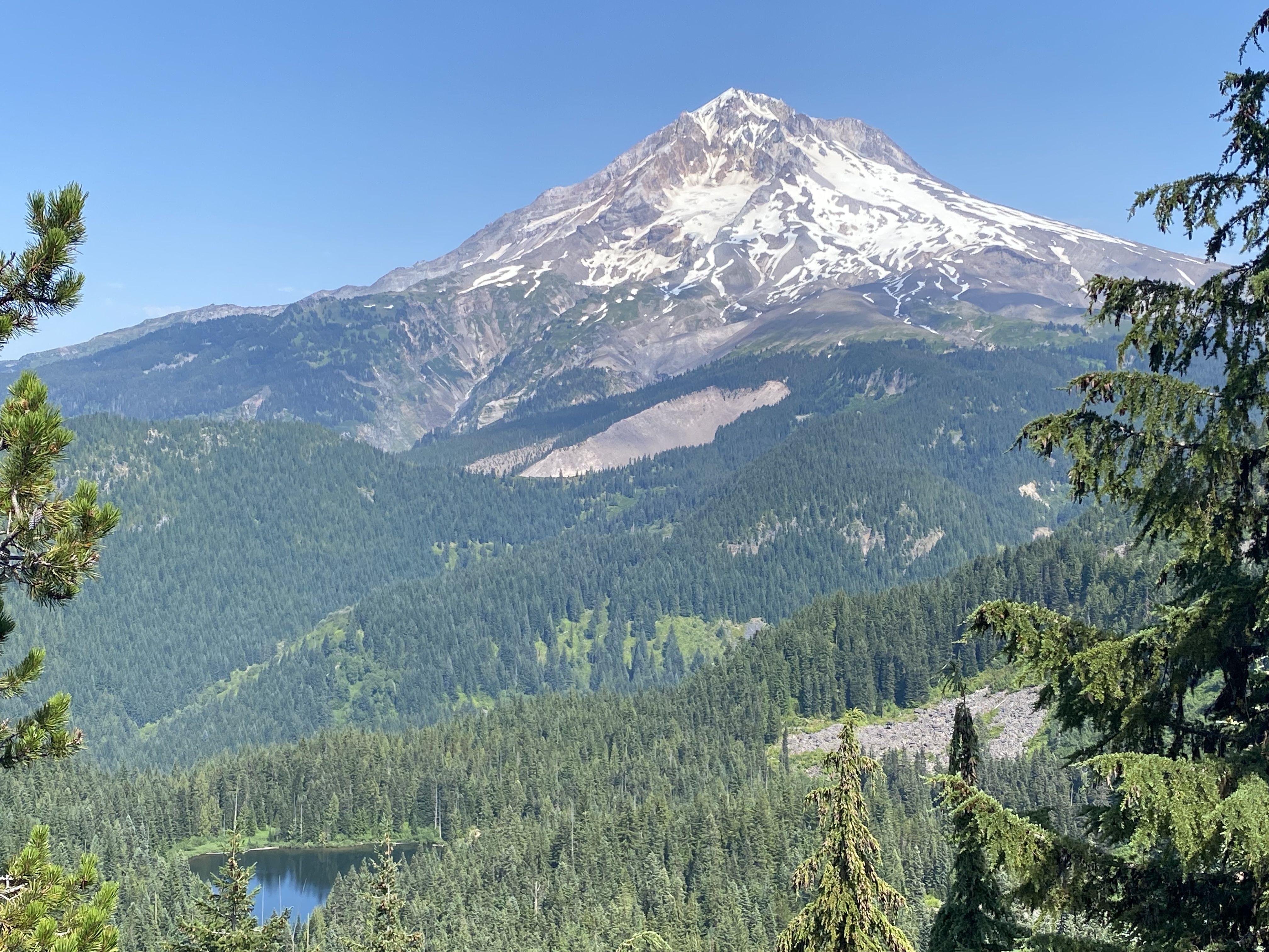 A wide image shows a small forested lake in the foreground with the summit of Mount Hood looming in the background.