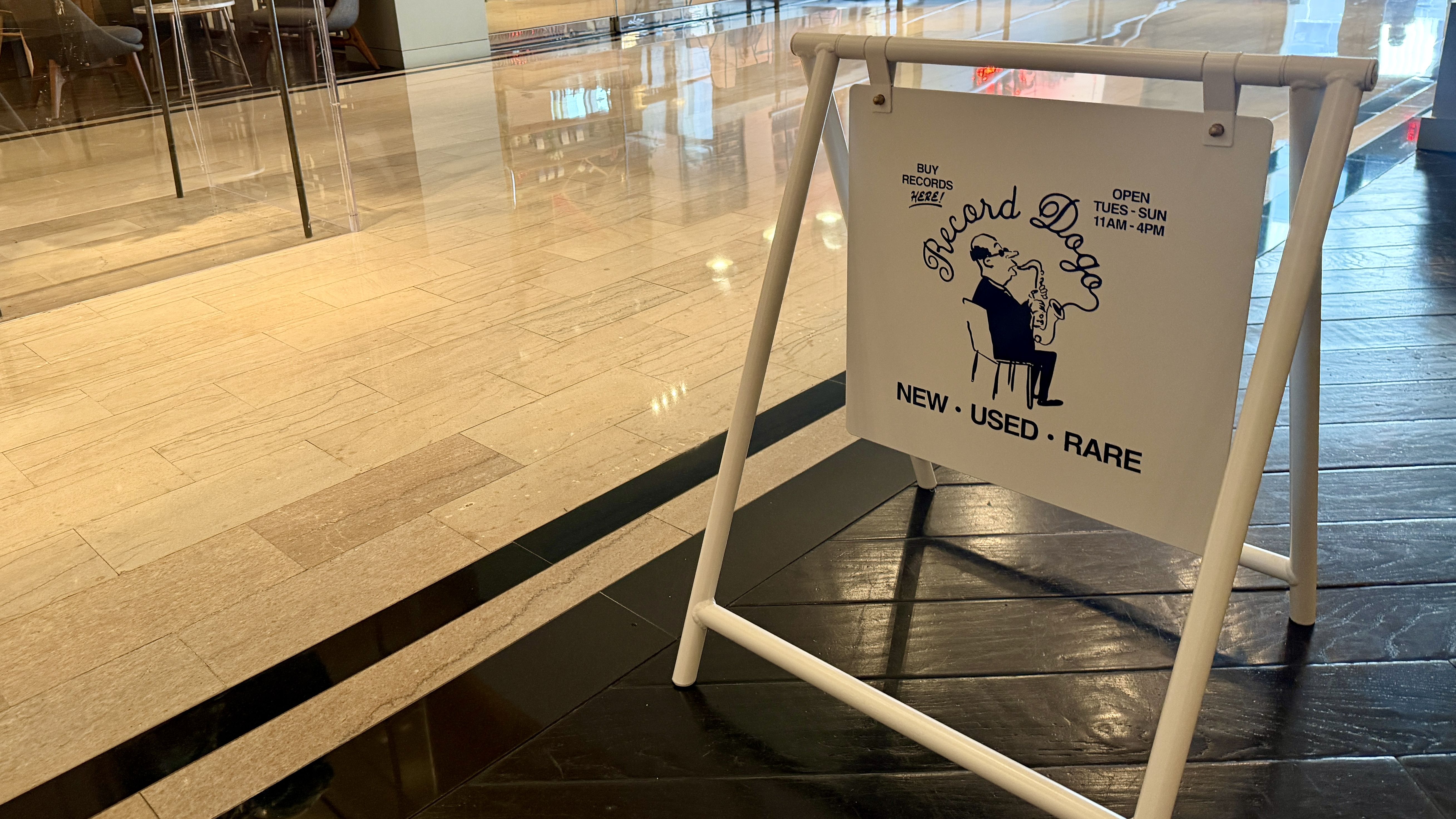Interior mall scene with a white A-frame sign showing a cartoon man in a suit playing a saxophone; text reads Buy Records Here, Open Tue-Sun 11am-4pm, New Used Rare.