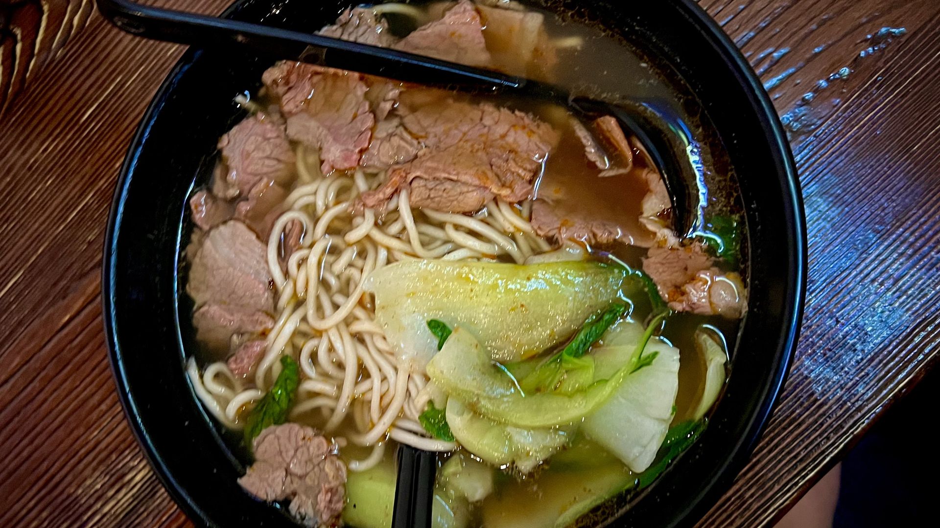 Black bowl of beef noodle soup with sliced beef, green leafy vegetables, and broth on a wooden table with black chopsticks and a spoon inside the bowl.