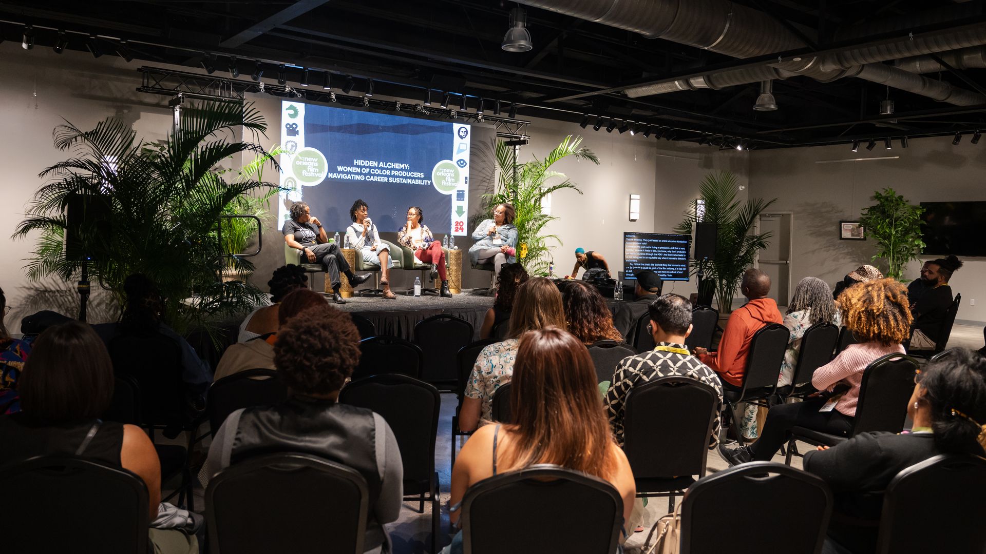 Panel discussion with four women seated on stage at New Orleans Film Festival, audience watching, indoor setting with plants and screen displaying topic.