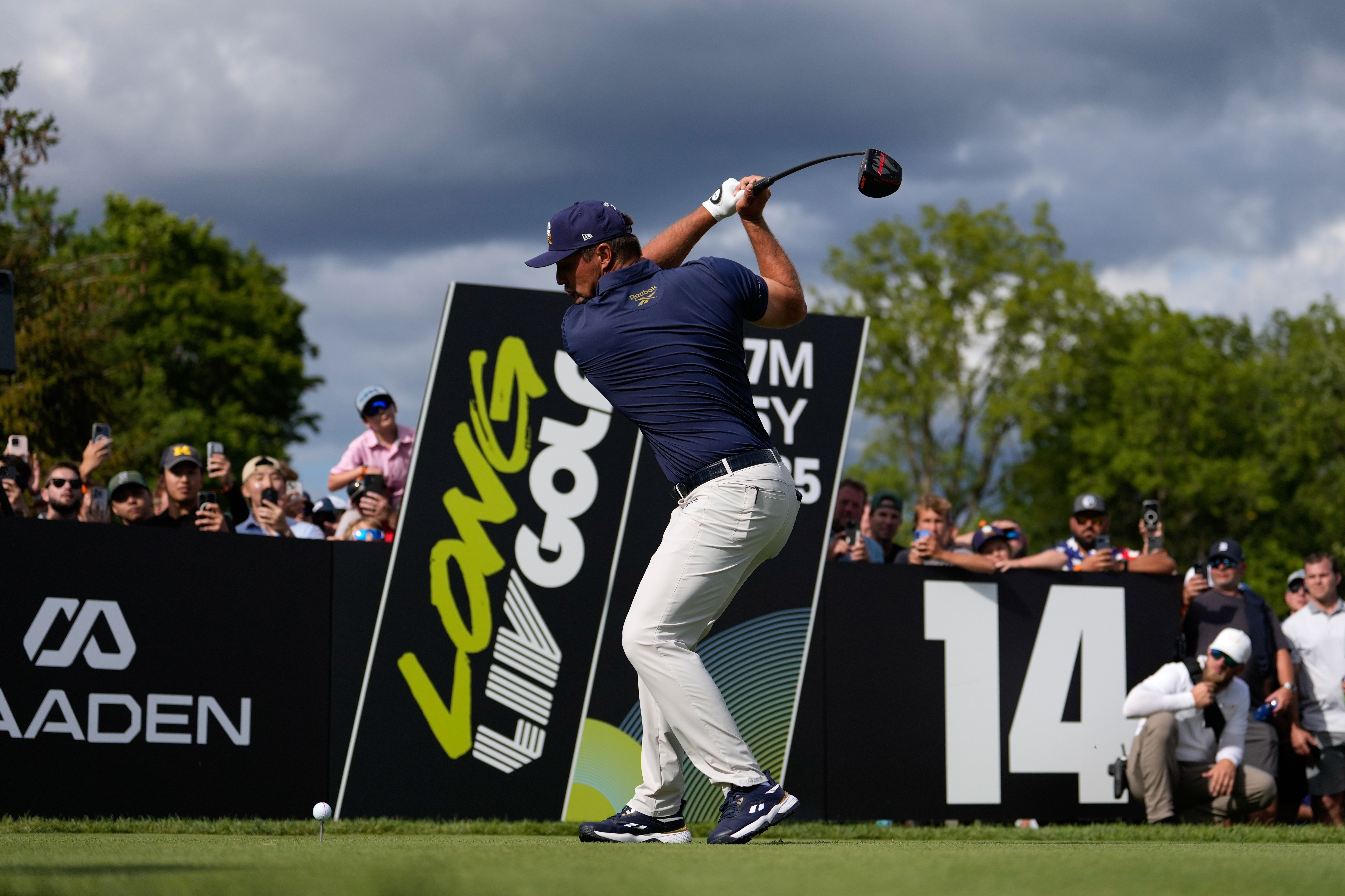 Golfer in navy shirt and white pants swings club on tee with crowd watching behind large sign showing 14 and "Laws Golf" under cloudy sky.