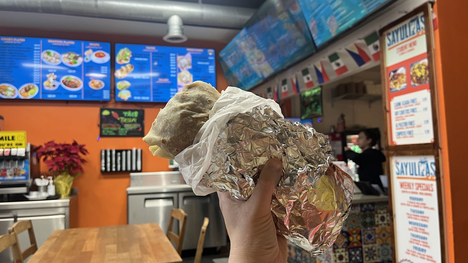 Hand holding a wrapped burrito inside a Mexican restaurant with orange walls, wooden tables, colorful tiled counter, and blue menu boards overhead.